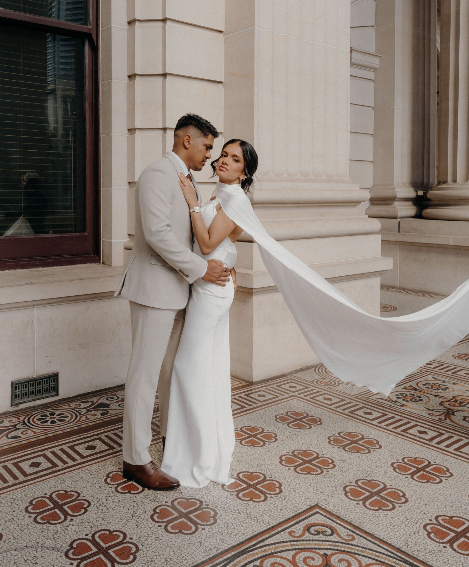 Newlywed couple in white attire dancing together on ornate tiled floor of historic building with flowing veil.
