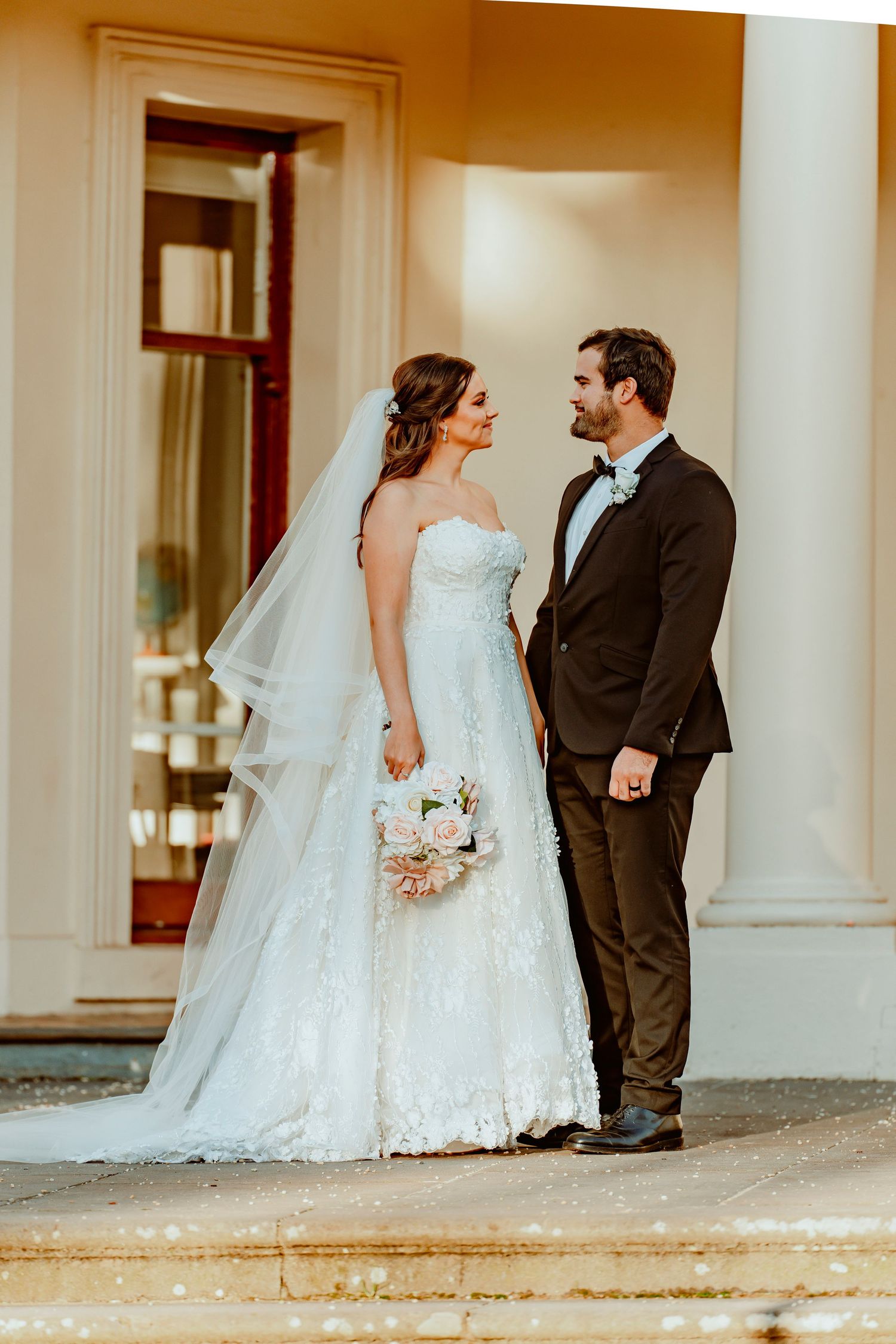 A bride in a white strapless gown and groom in black tuxedo share a candid moment outside an elegant venue.