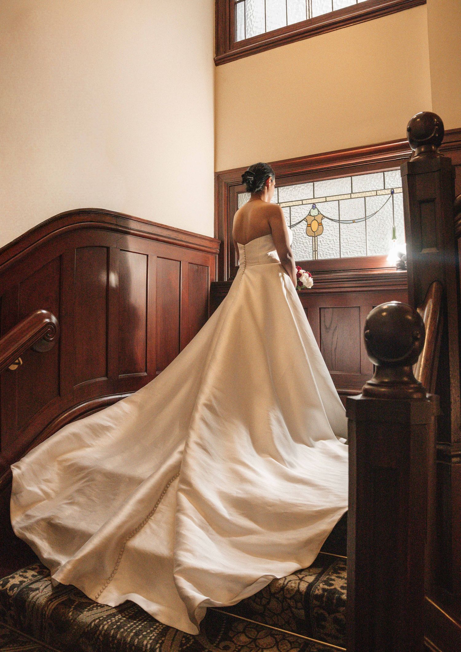 Bride in elegant white ball gown with long train stands on ornate wooden staircase near stained glass window.