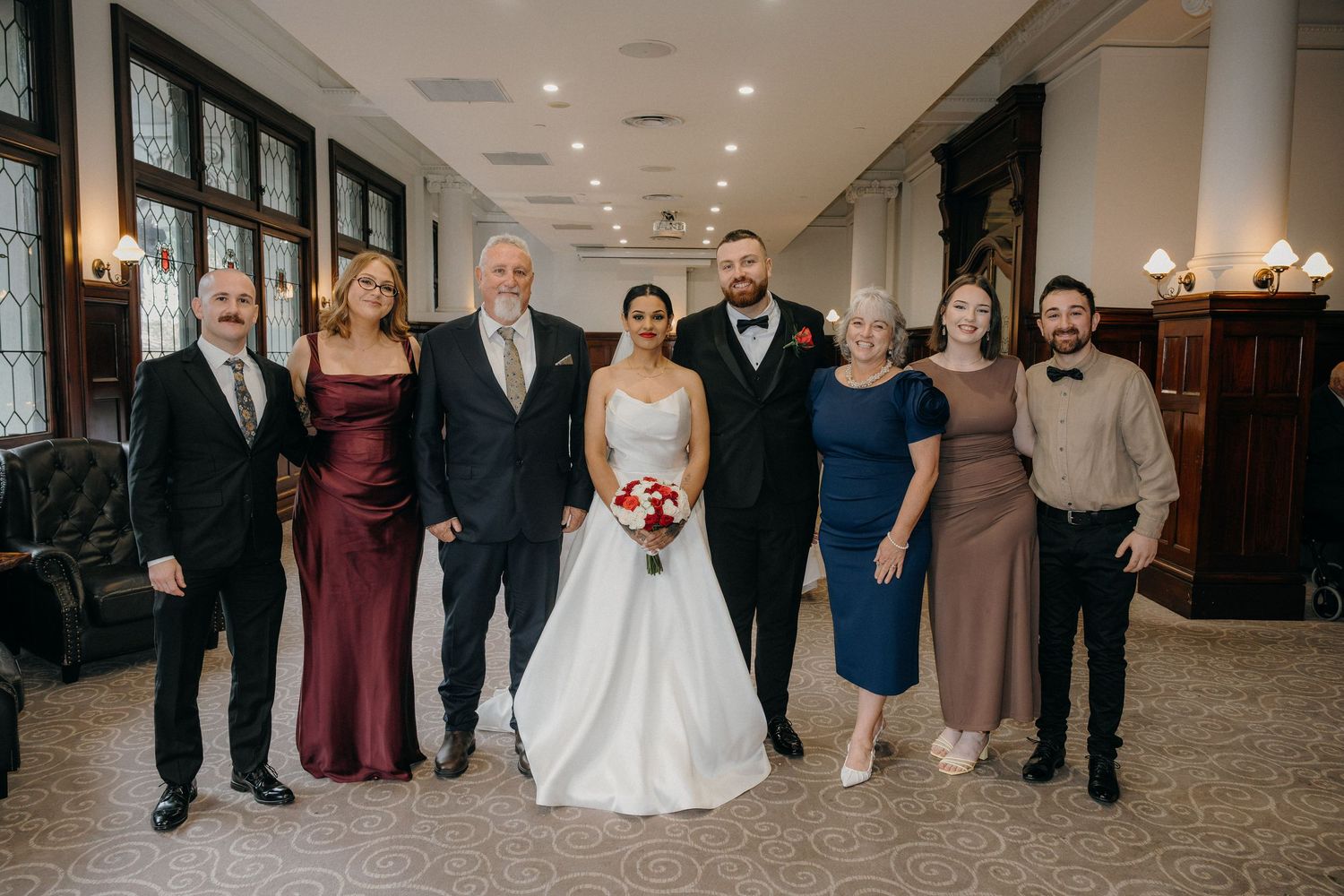 Wedding party poses indoors, bride in white gown holding bouquet, groom and six formally dressed family members smiling together.
