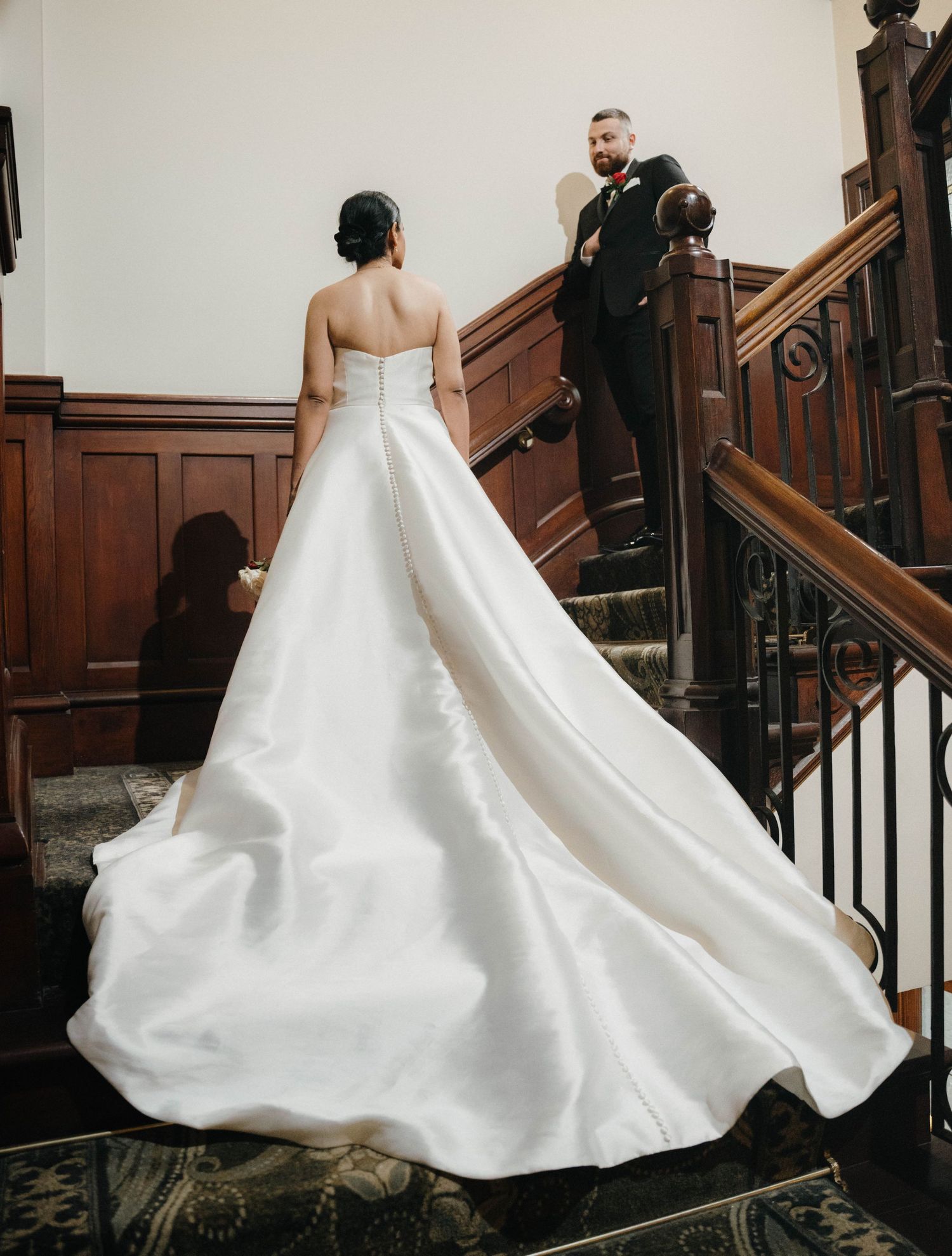 Bride in elegant strapless white ball gown with long train on staircase, groom in black suit above her.