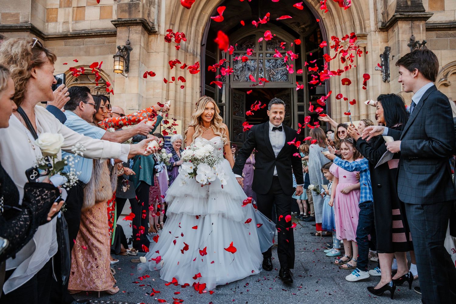 Wedding guests toss red rose petals over the newlyweds as they exit a historic stone church after their ceremony.