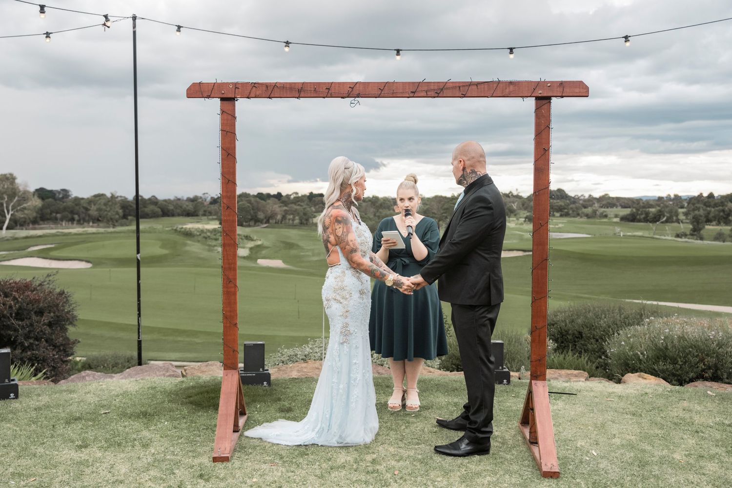 A wedding ceremony takes place under a wooden arch structure on a green golf course with string lights overhead.