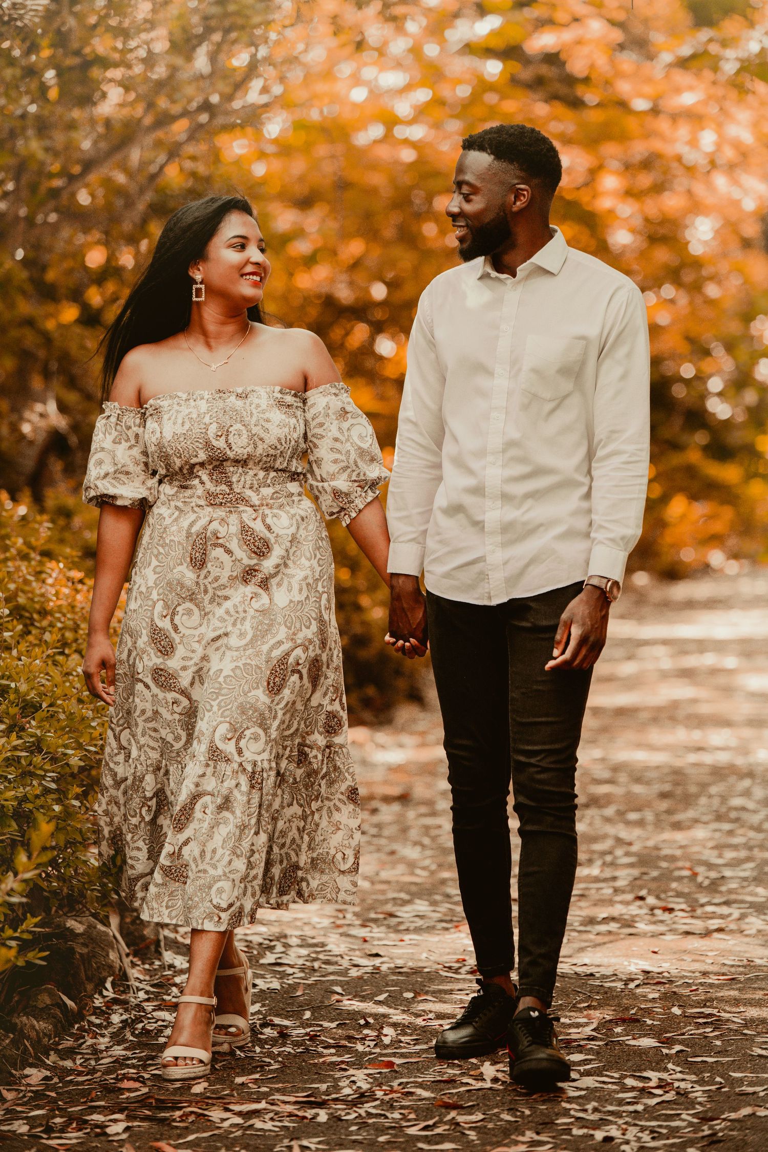 A couple walks together on a leaf-covered path surrounded by vibrant autumn foliage in golden orange hues.