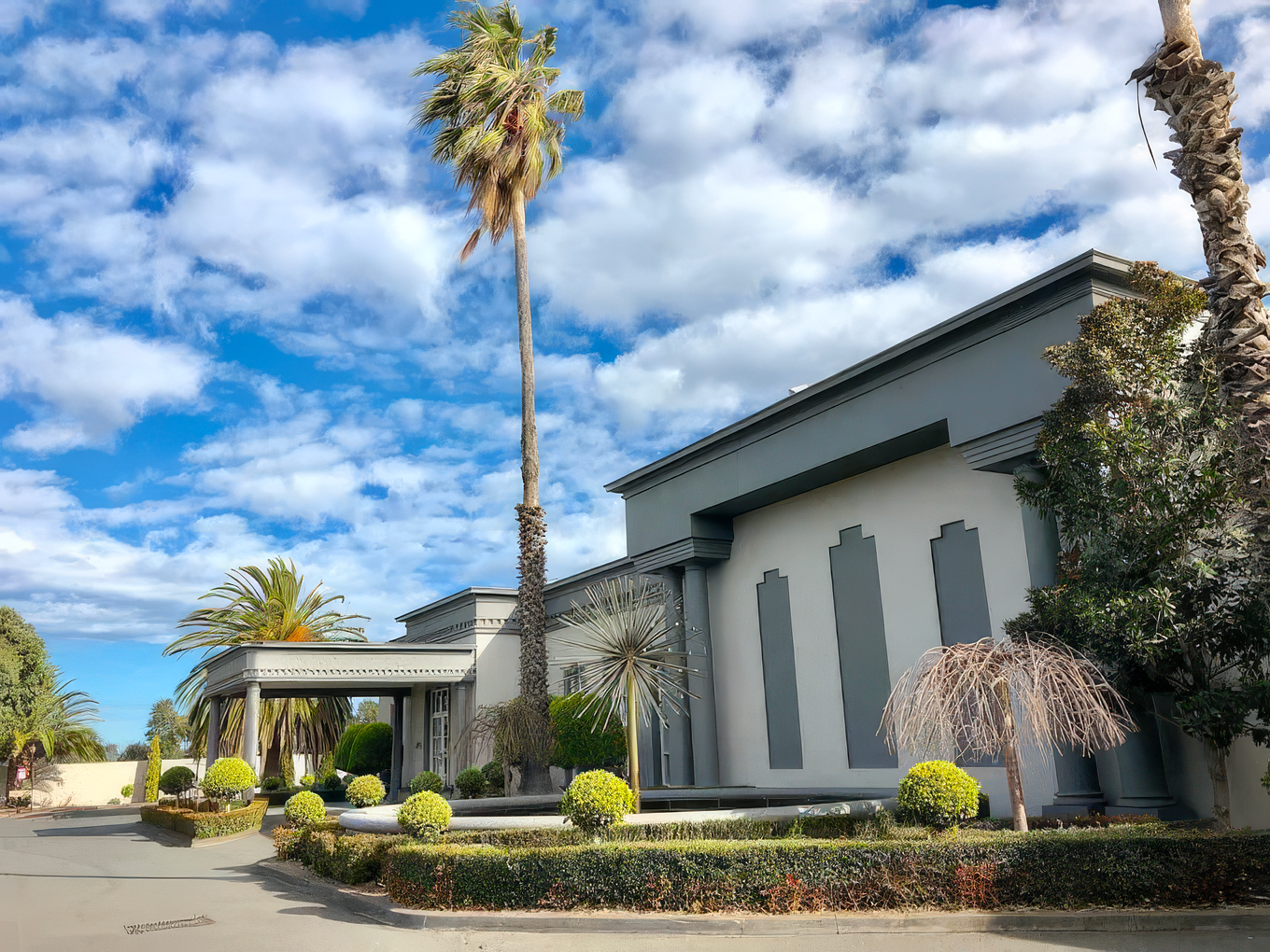 Modern church building exterior with palm trees and landscaping against bright blue sky with white clouds.