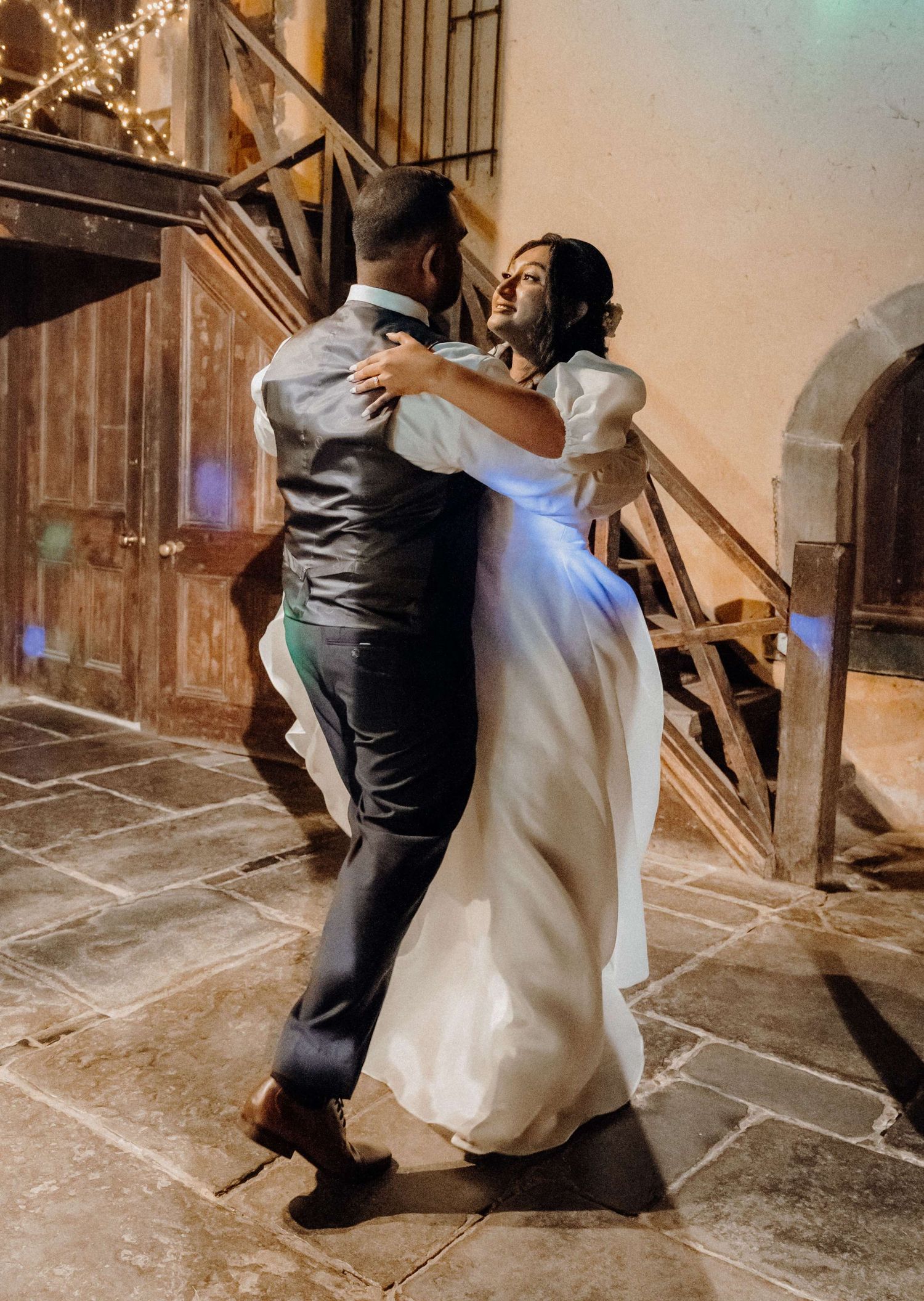 A dog in a tuxedo affectionately hugs a bride in her white wedding dress on a concrete floor.