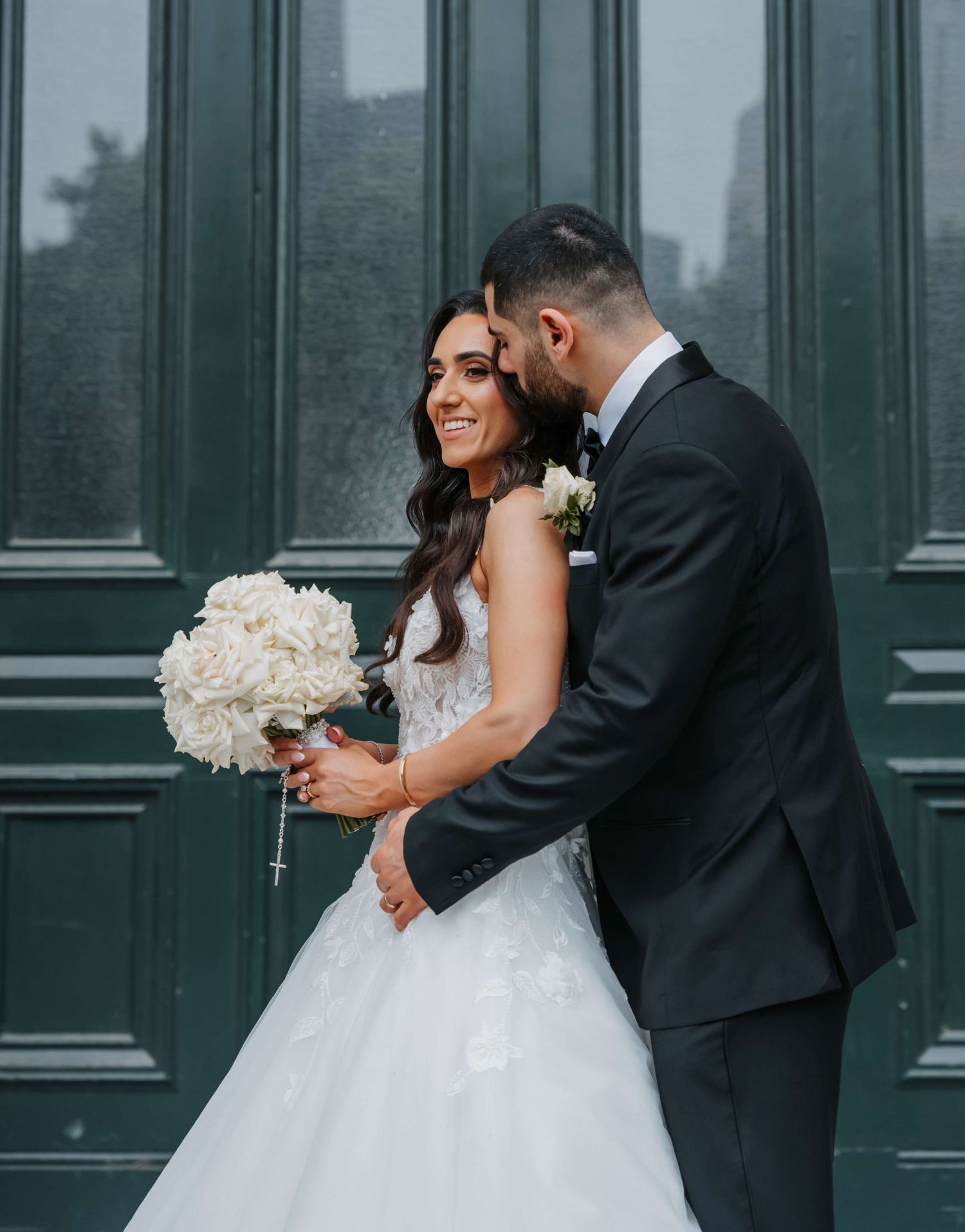 Joyful wedding couple shares an intimate moment against green doors while holding a white peony bouquet.