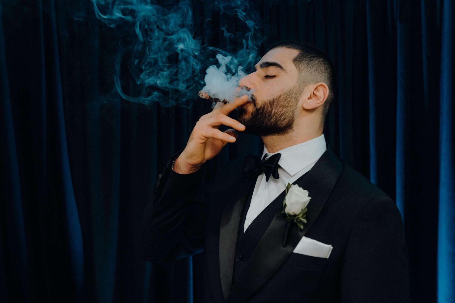 A person in a formal black tuxedo exhales smoke from a cigarette against a dark blue backdrop.