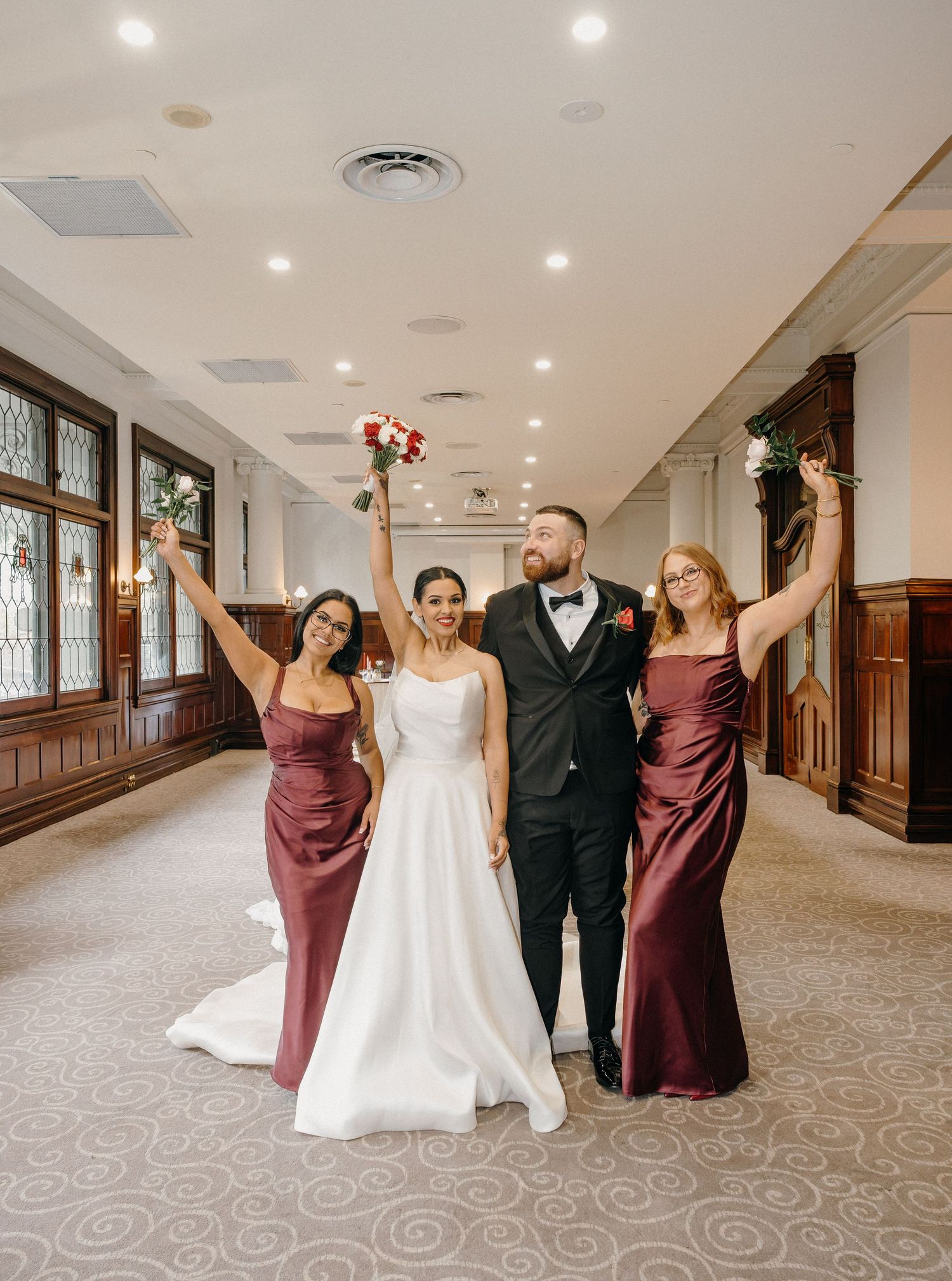 Bride and groom celebrate with two bridesmaids in burgundy gowns, raising bouquets in an elegant wedding venue hallway.