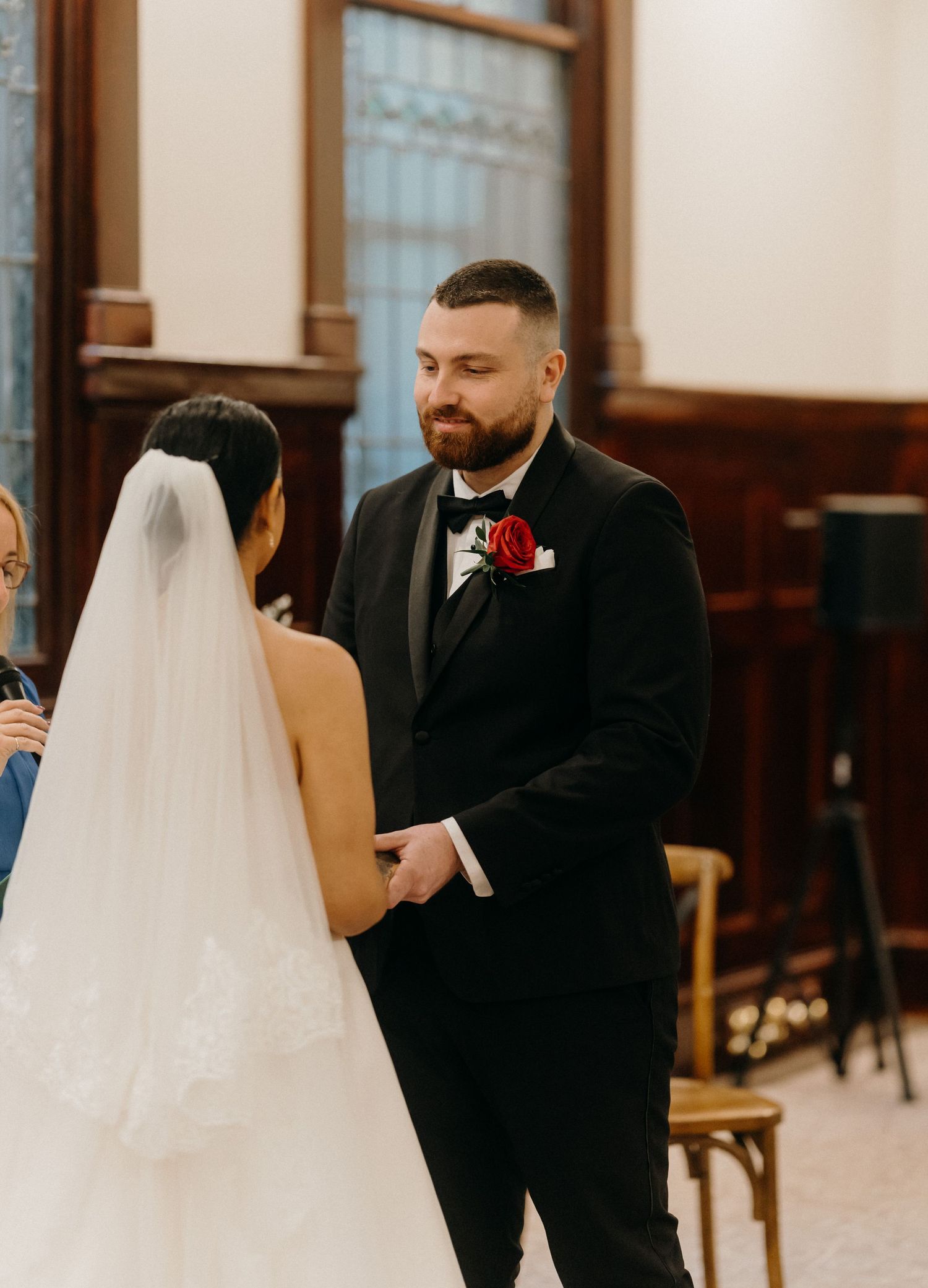 Groom in black tuxedo with red rose boutonniere holds bride's hands during indoor wedding ceremony.
