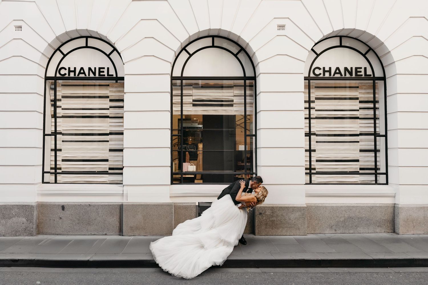 A white wedding dress flowing on the sidewalk outside a Chanel storefront with three arched windows and black metal signage.