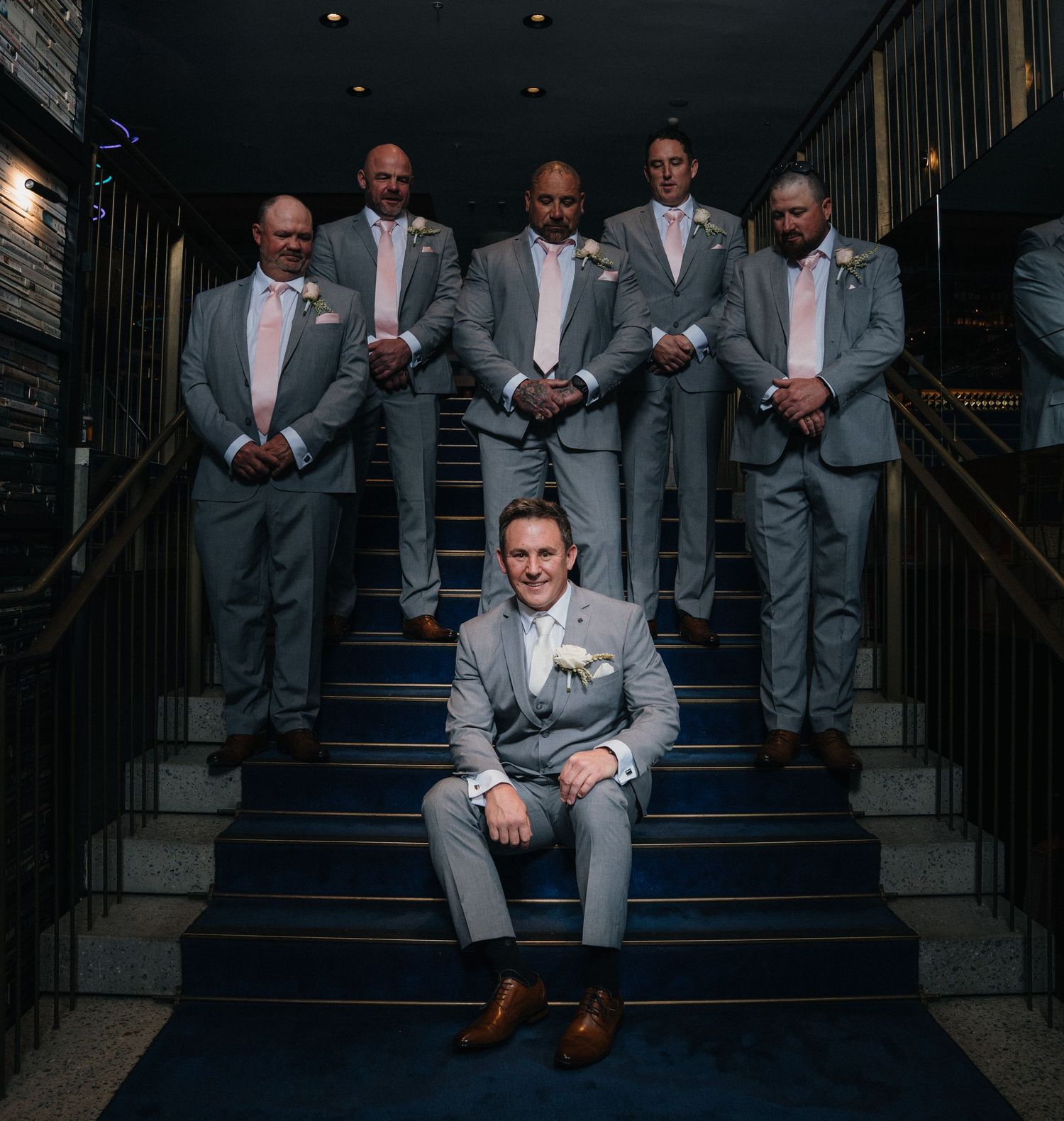 Groomsmen in matching gray suits pose on dark staircase for formal wedding photos.