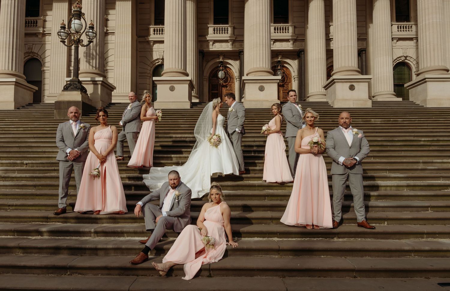 Wedding party in formal attire poses on grand stone steps of historic building with classical columns.