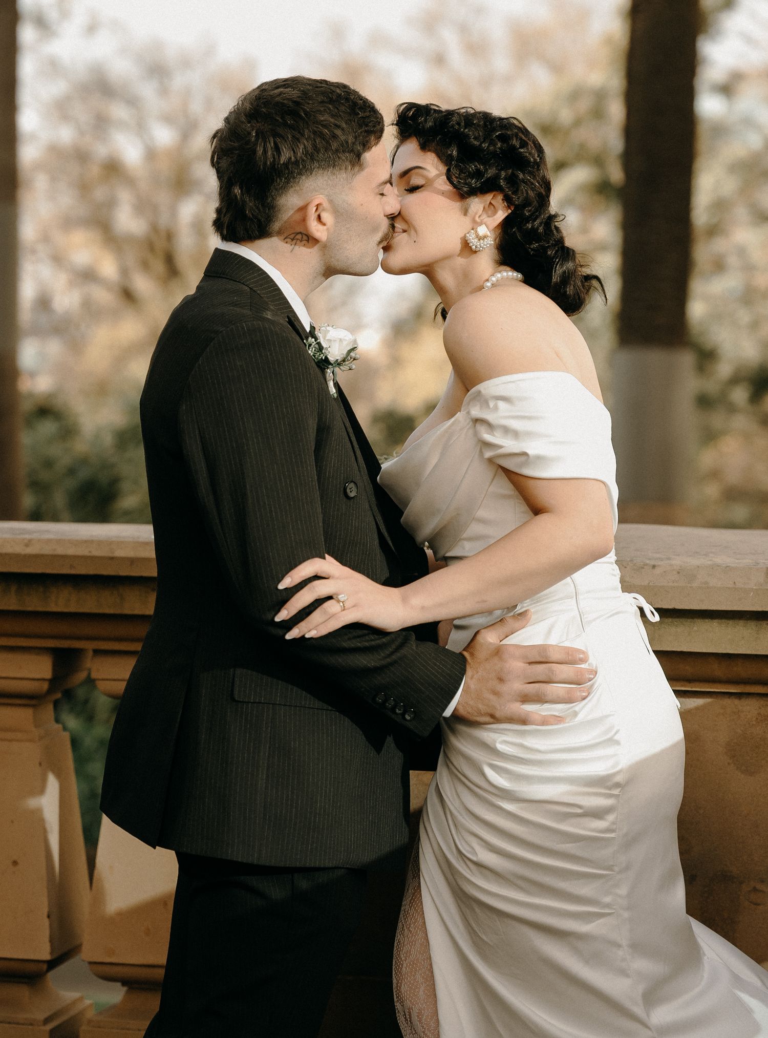 A couple shares a romantic kiss in wedding attire, bride in white off-shoulder gown, groom in dark suit.