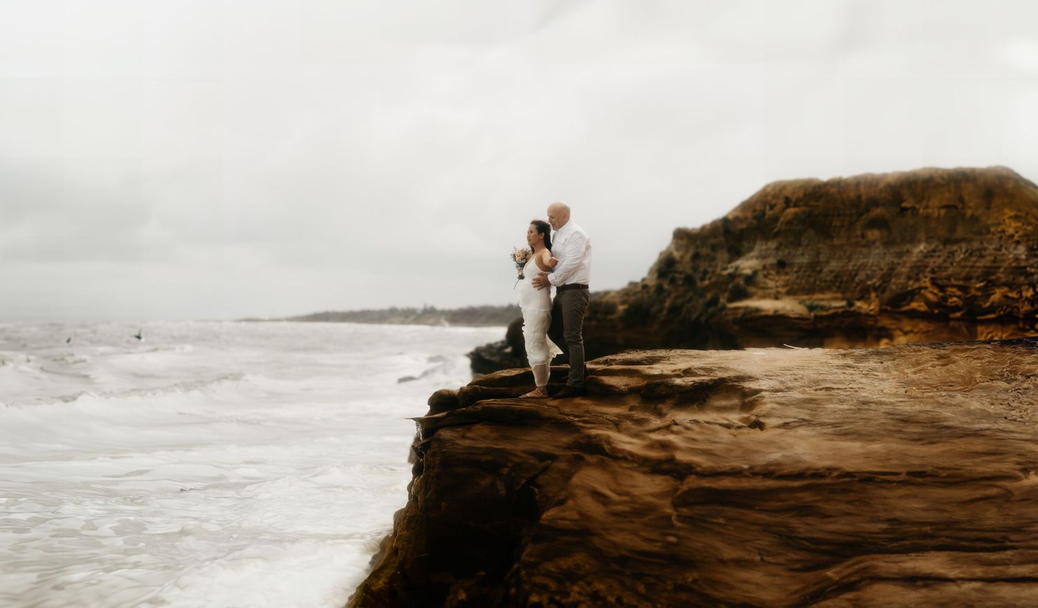 A person sits on a rocky cliff overlooking a misty coastline with crashing waves below.