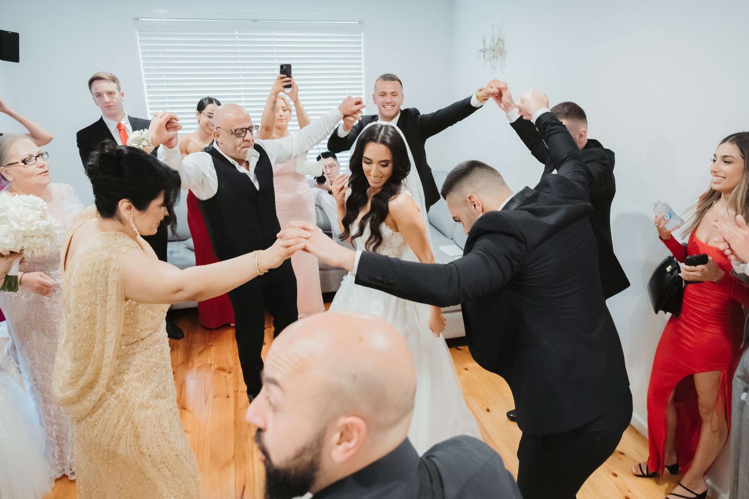 A festive crowd cheers and raises their arms during a celebratory dance moment at an indoor wedding reception.