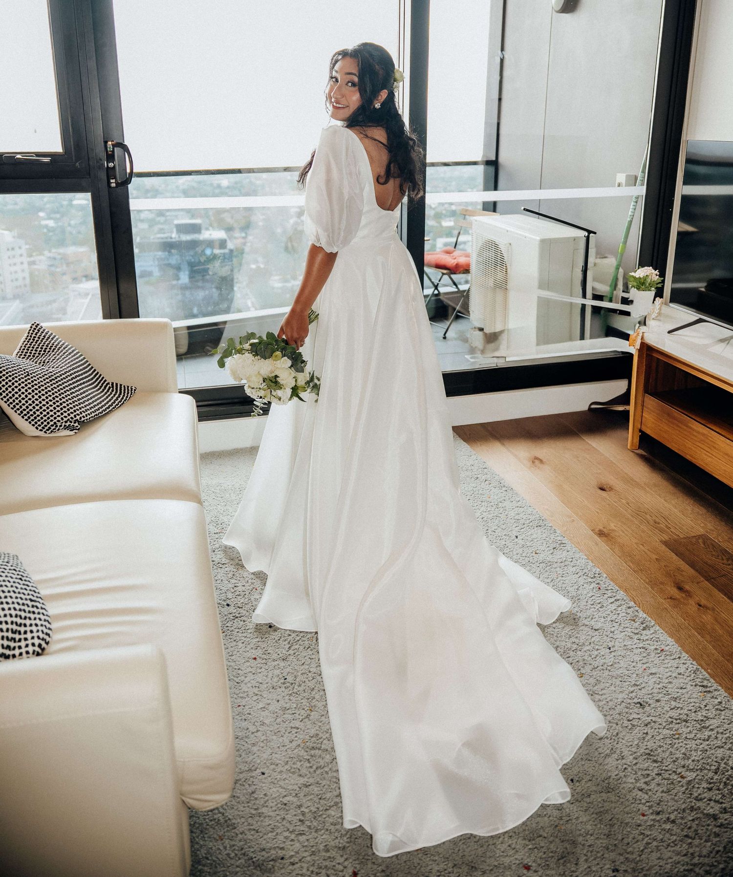 Woman in flowing white wedding dress standing by window in modern apartment with city views and hardwood floors.