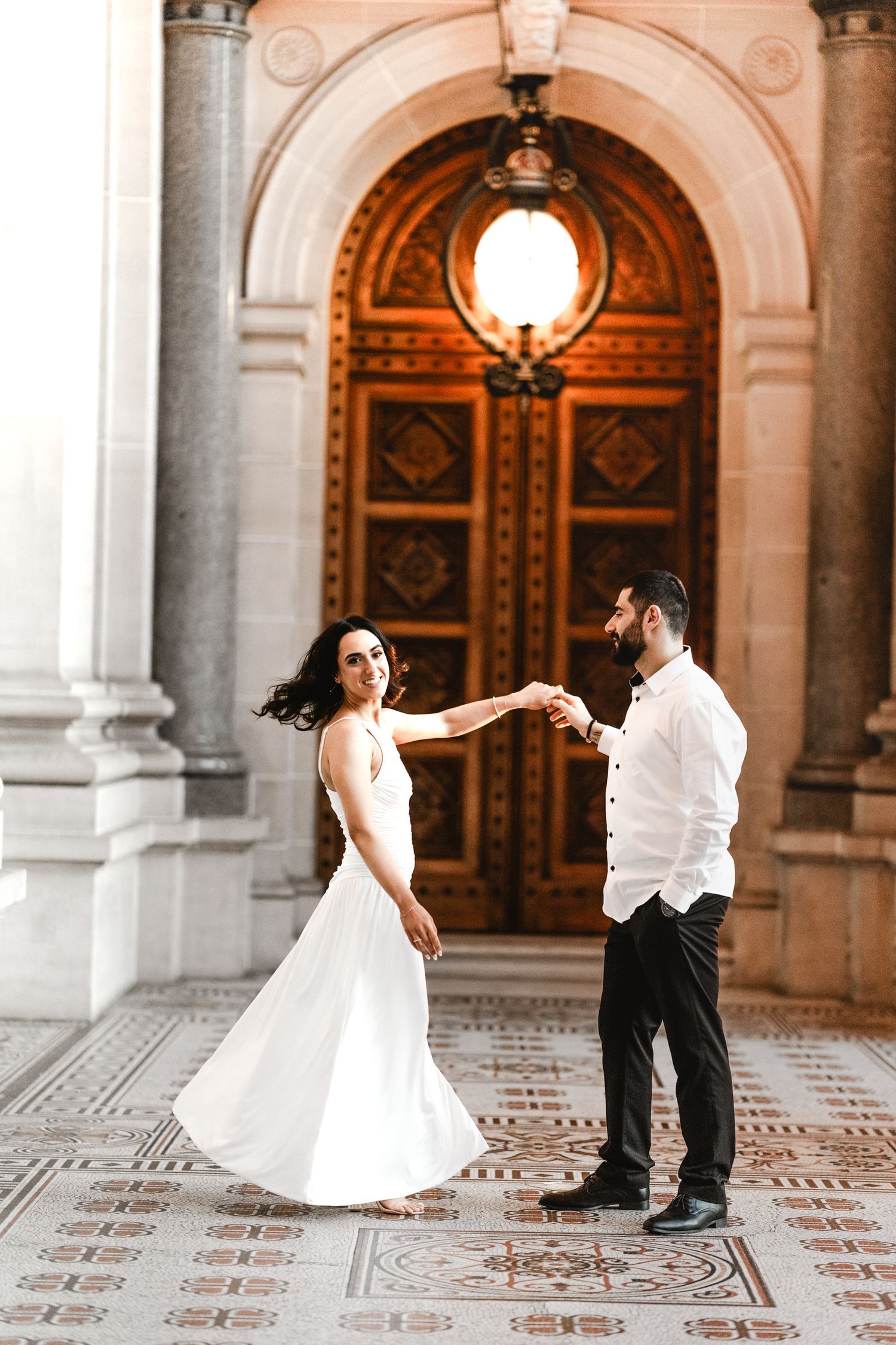 A couple dances together on an ornate tiled floor beneath a grand arched doorway with vintage lighting.