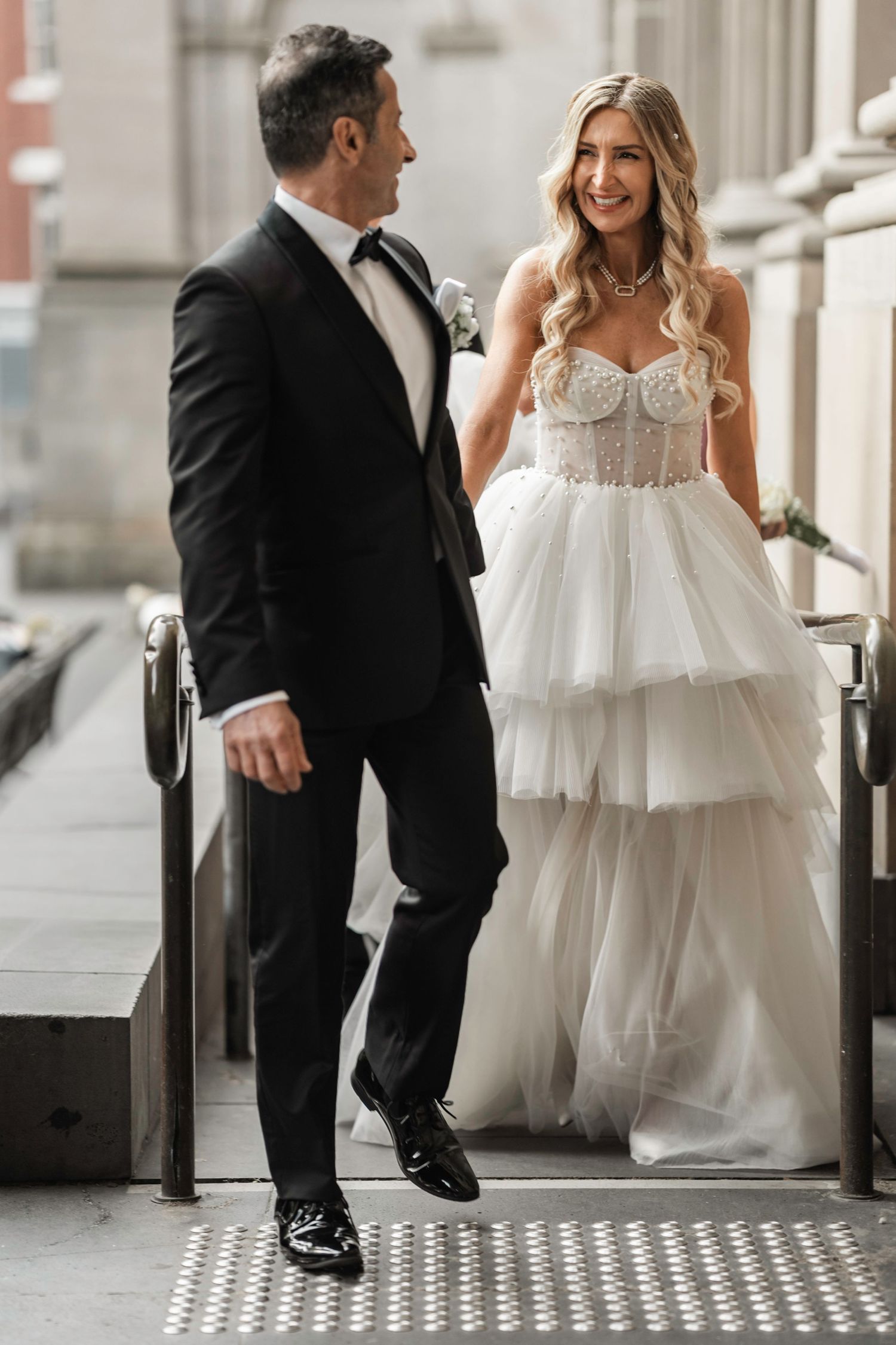 A bride in a flowing white dress and groom in black suit dance playfully on a patterned floor in an elegant venue.
