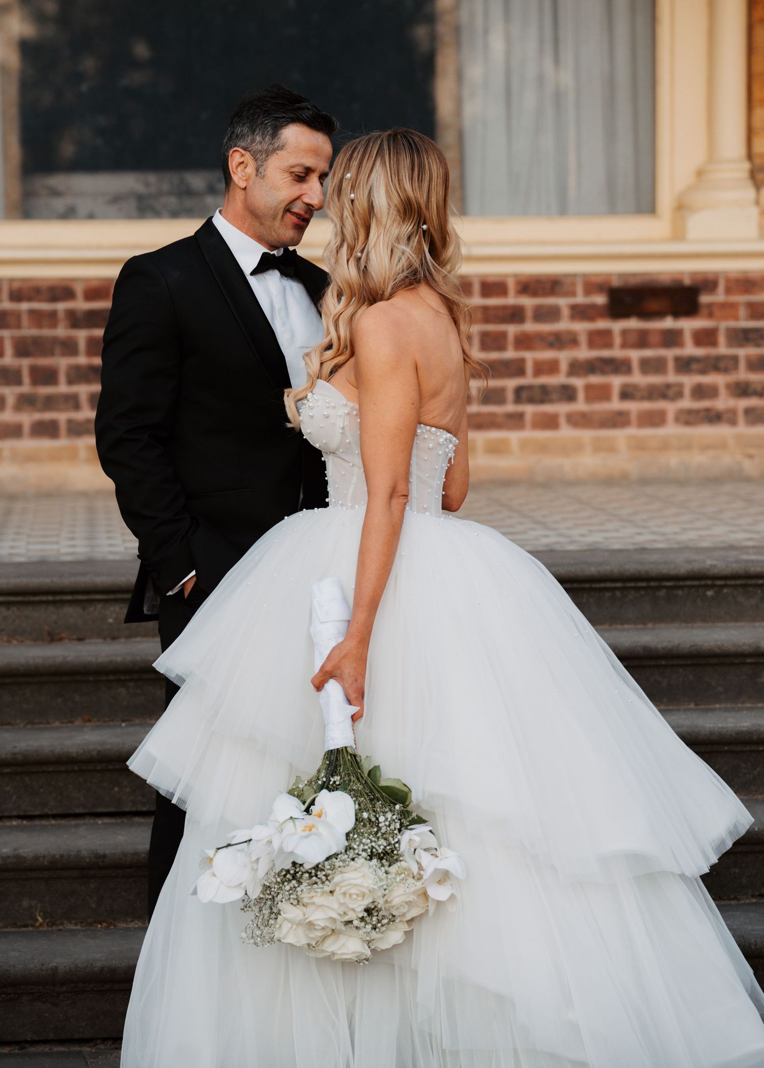 A romantic wedding couple embraces on stone steps, with the bride wearing a layered tulle ballgown and holding white flowers.