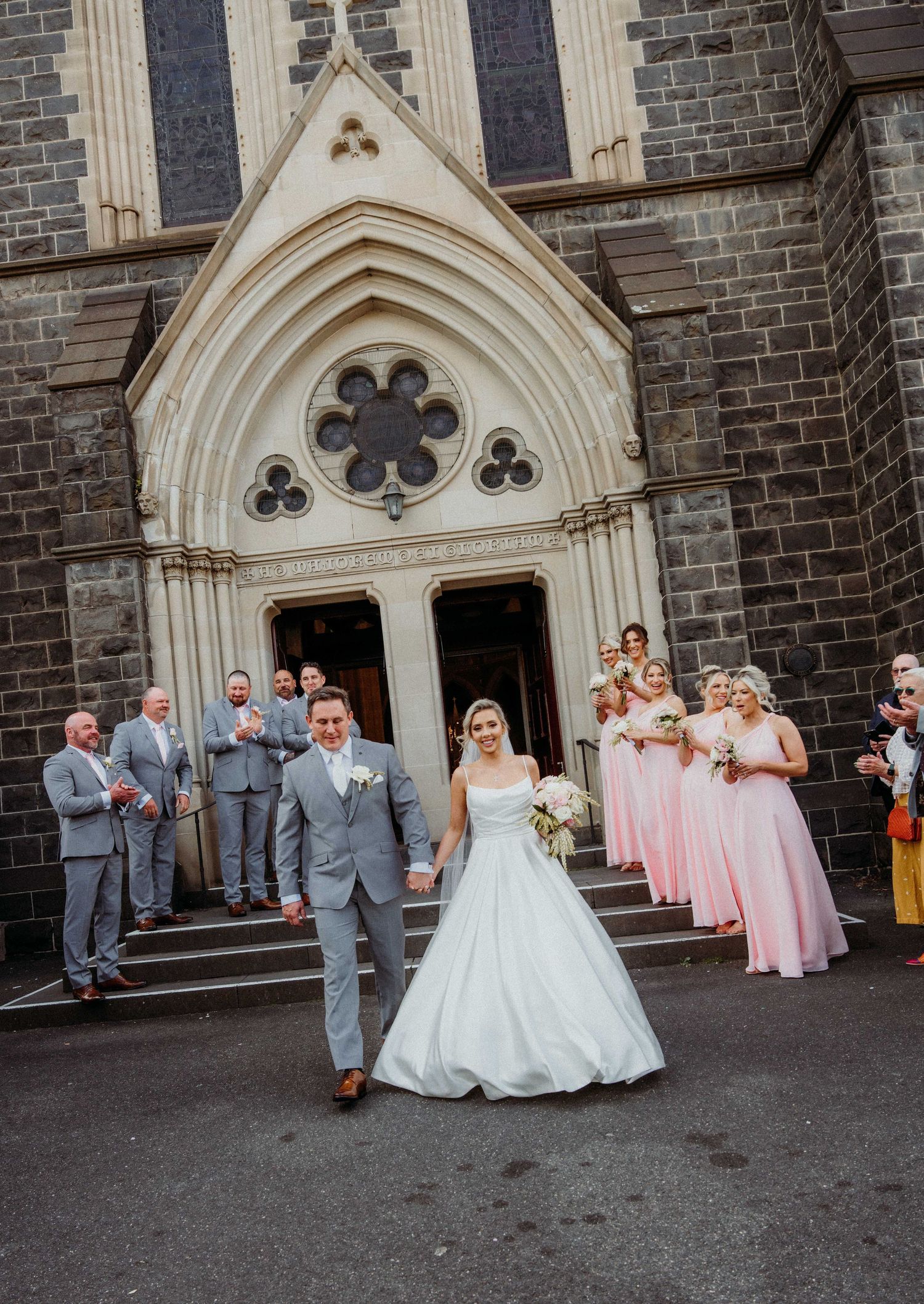 Wedding party poses in front of a gothic stone church with pink bridesmaids dresses and gray suits.