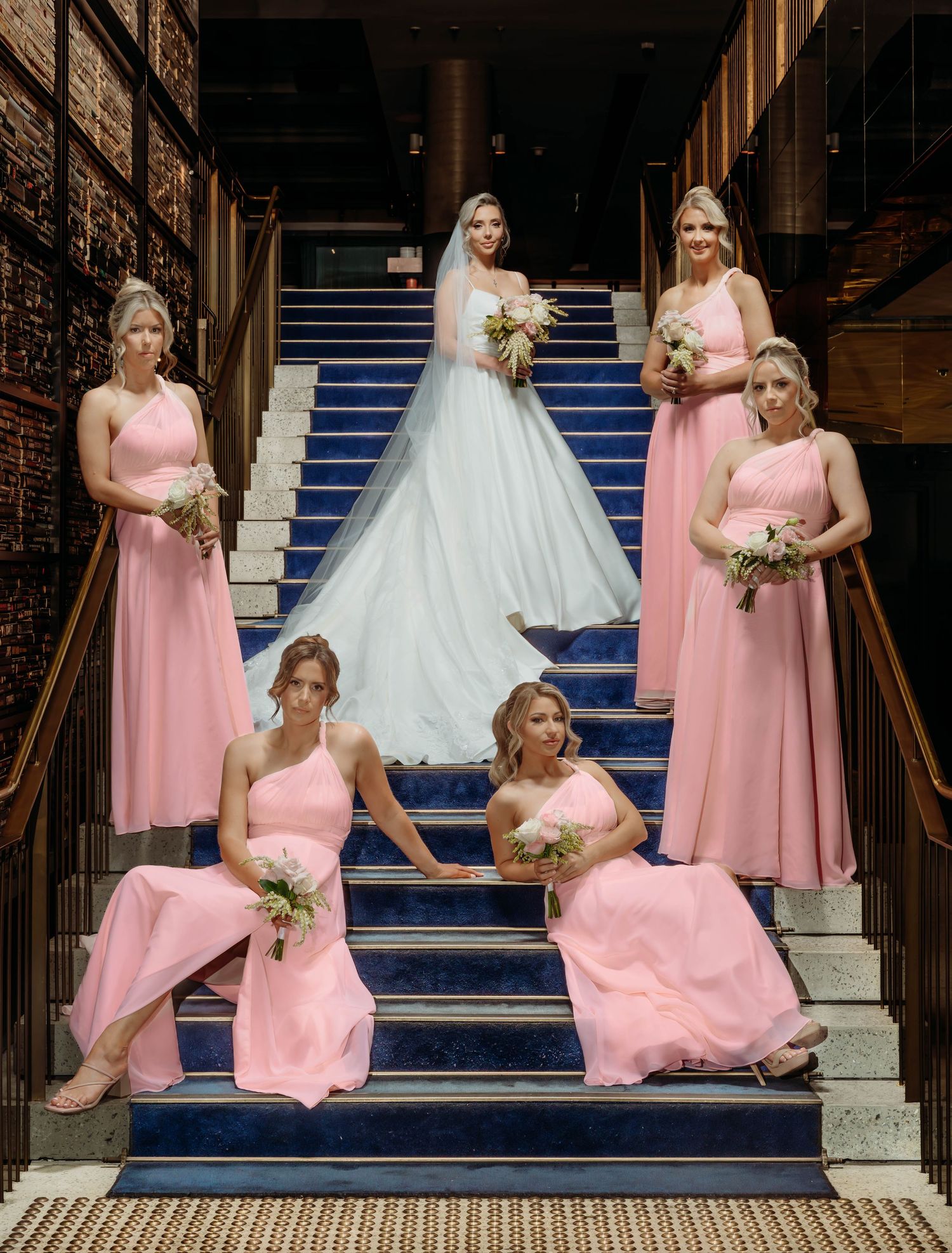 A bridal party in pink dresses poses playfully on elegant blue carpeted stairs for wedding photos.