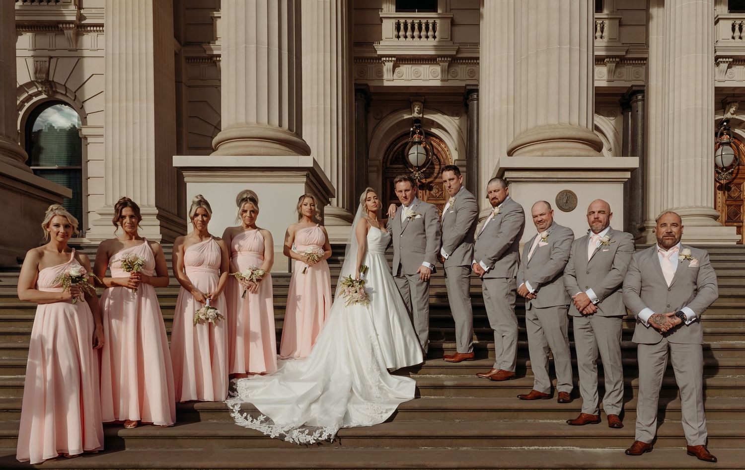 Wedding party members sit casually on grand stone steps of a historic building with classical columns.