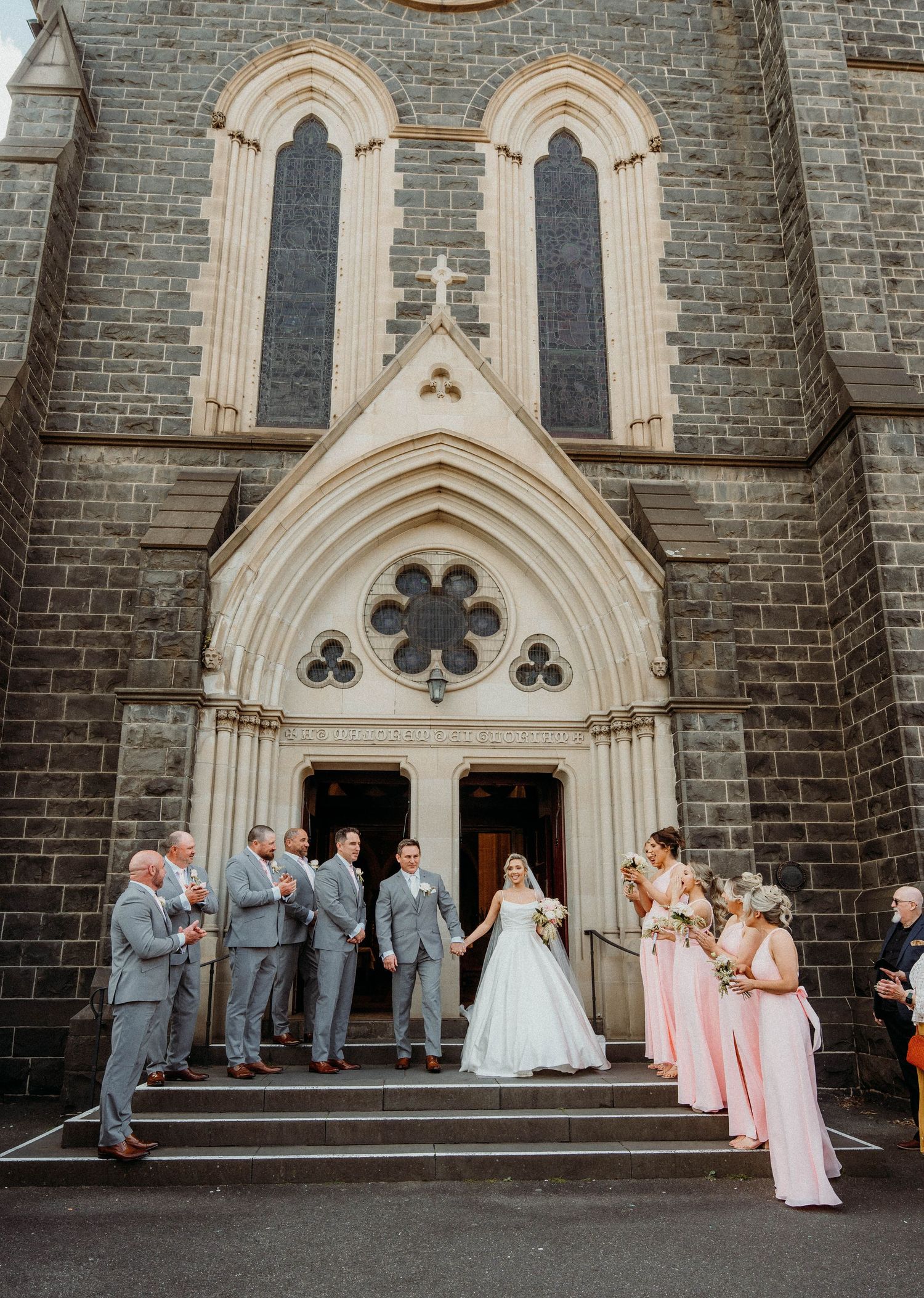 Wedding party poses on church steps with bride in white gown and bridesmaids in pink dresses.