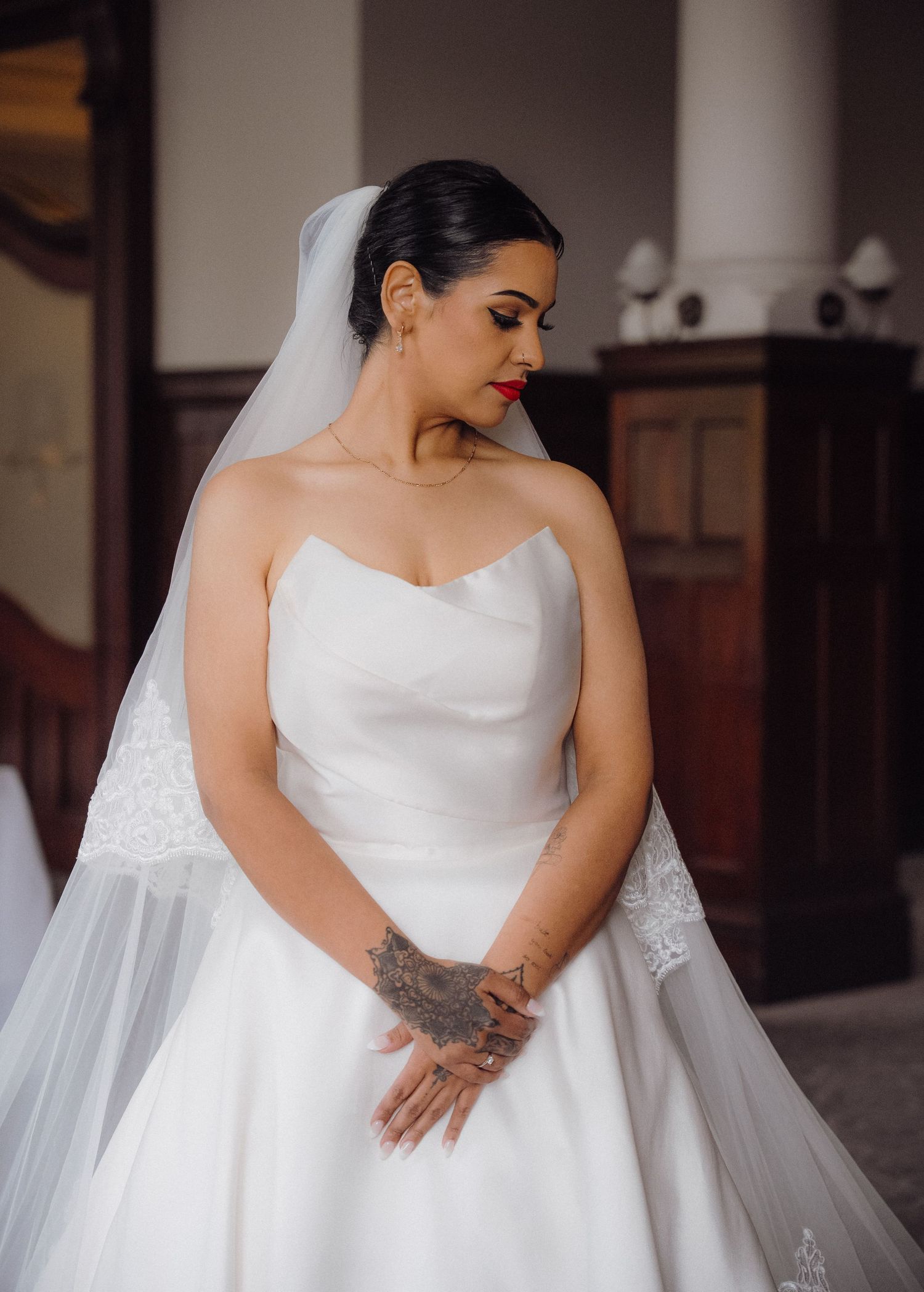 Bride in strapless white ball gown with lace veil and red lipstick, hands clasped, tattoo visible.