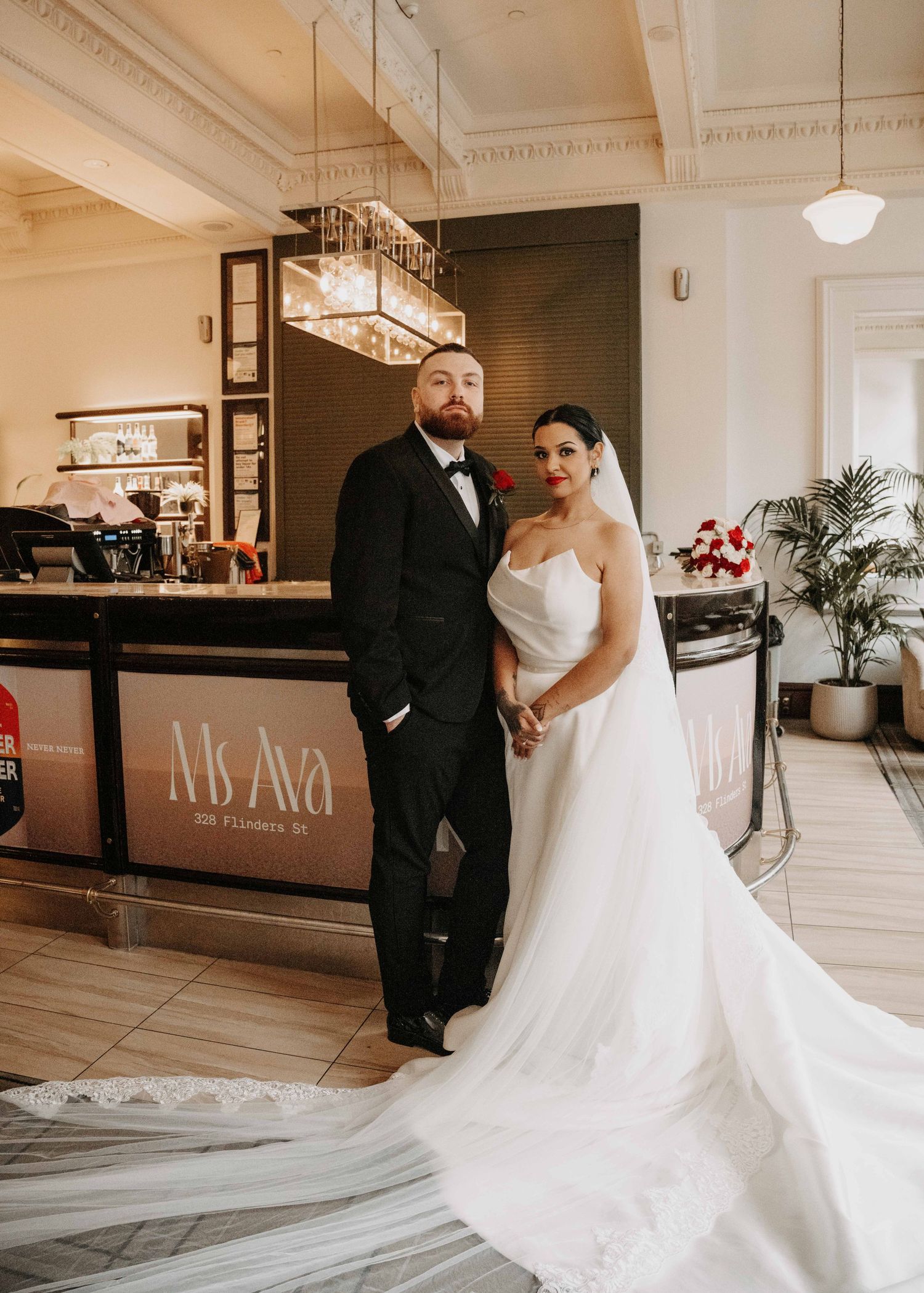 Bride in white strapless gown with long veil poses with groom in black tuxedo at Ms. Ava restaurant wedding venue.