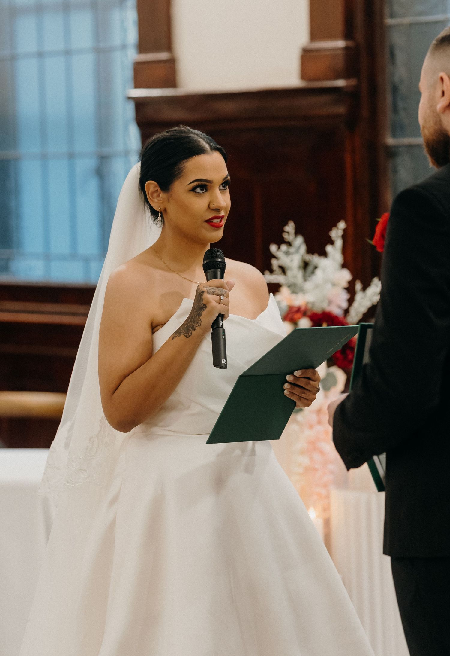 Bride in white strapless gown and veil reads vows into microphone at wedding ceremony altar.