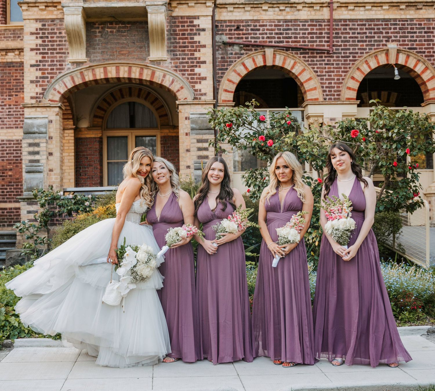 Wedding party in dusty purple mauve bridesmaid dresses poses outdoors against a brick building with floral archway.