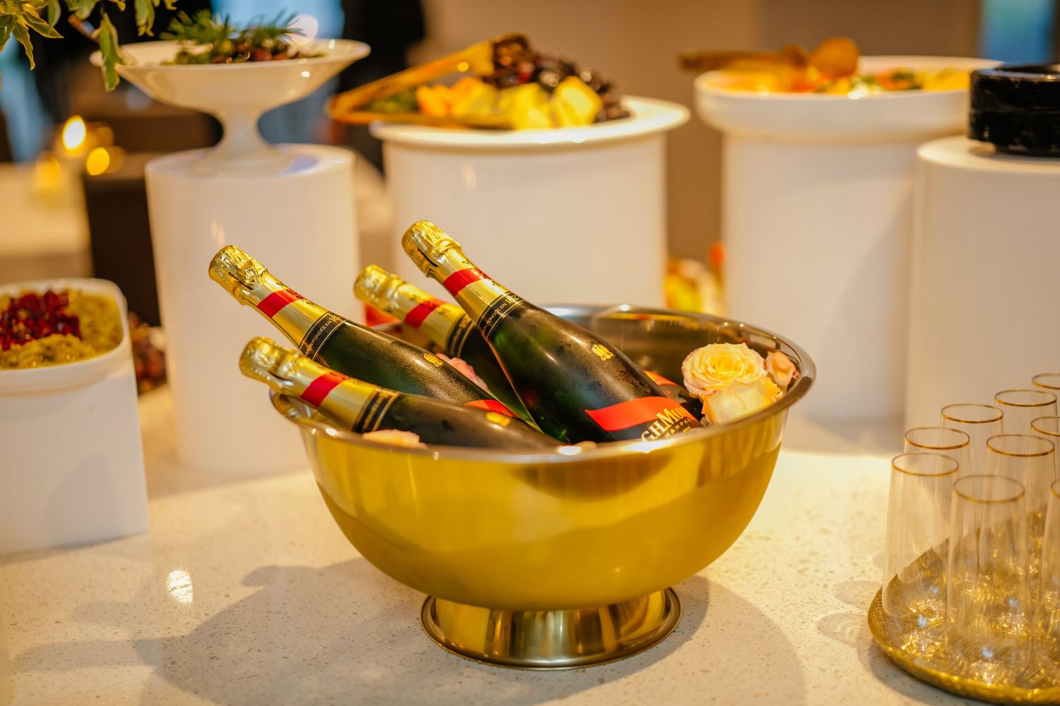 Assorted cigars displayed in elegant gold metallic bowls or vessels with clear glassware nearby on a white surface.