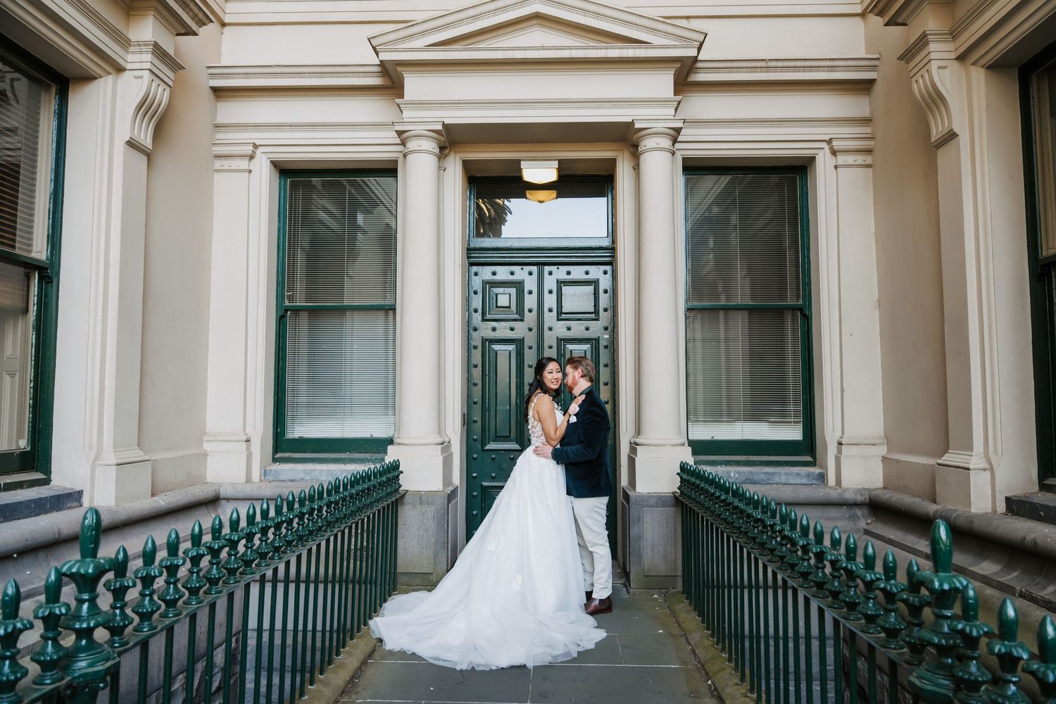 Bride and groom share an intimate moment on an elegant balcony with white columns and green railings.
