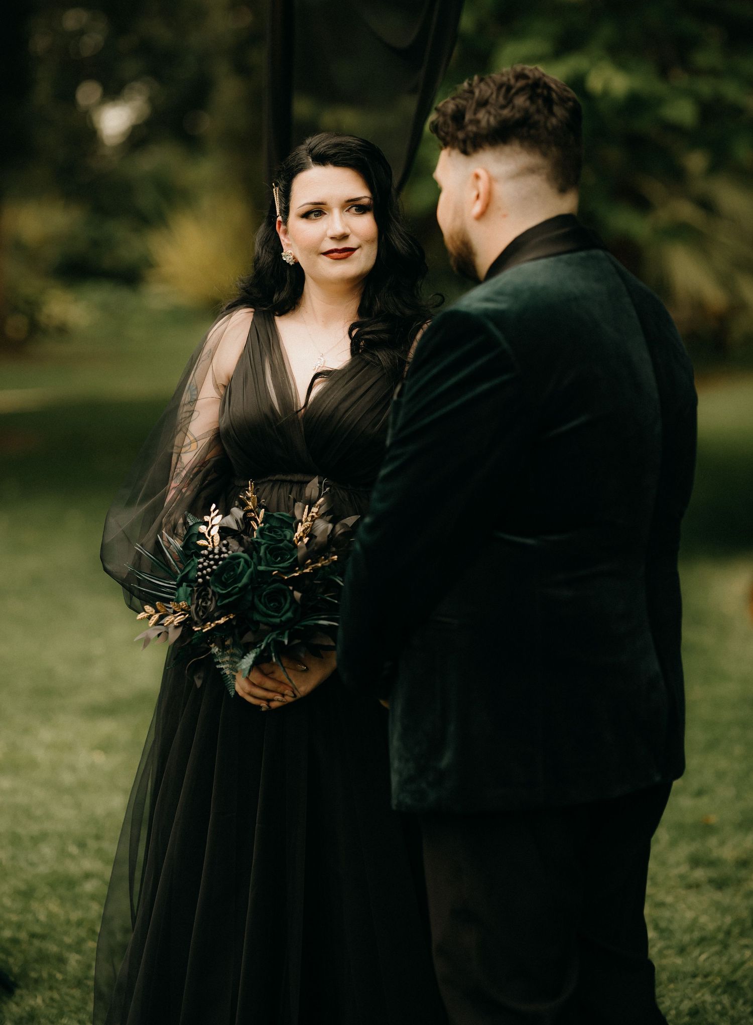 A couple in dark formal attire stands together outdoors during a moody wedding photoshoot with natural greenery backdrop.
