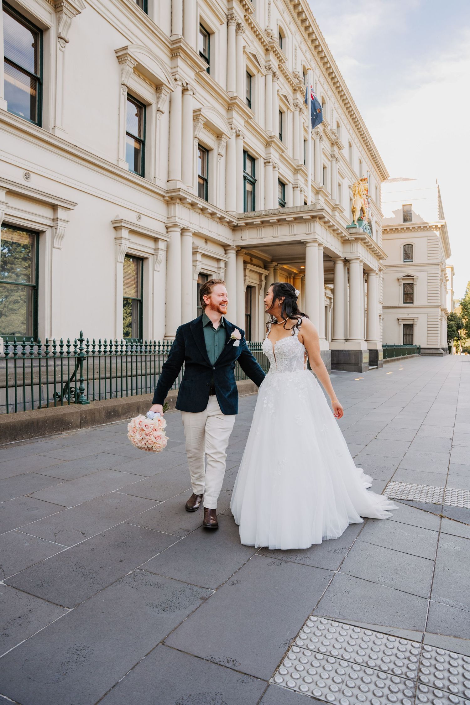 A newlywed couple strolls along a historic building exterior with elegant white stone architecture and ornate details.