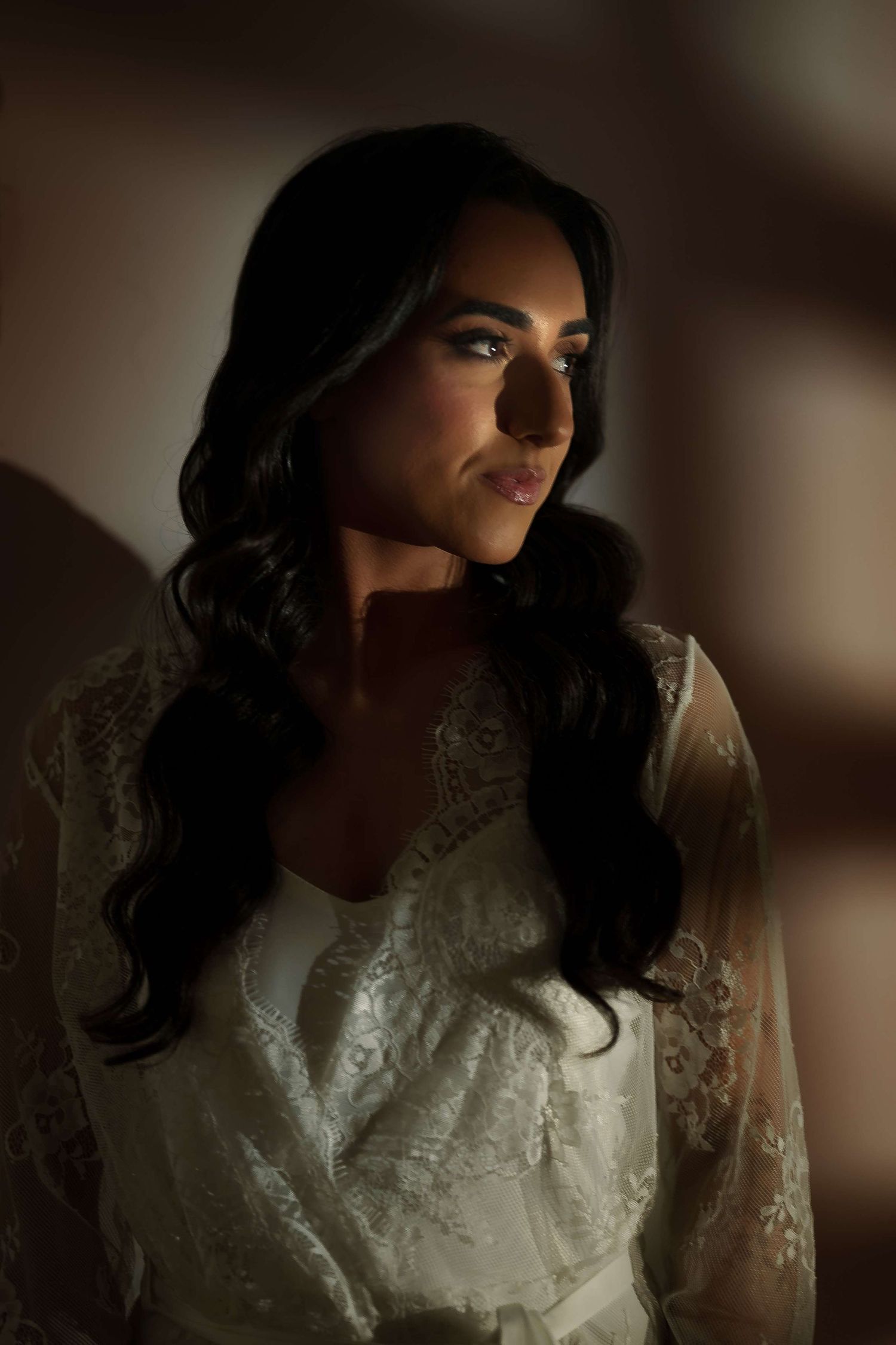 A bride in a white lace dress poses in soft moody lighting with dramatic shadows.