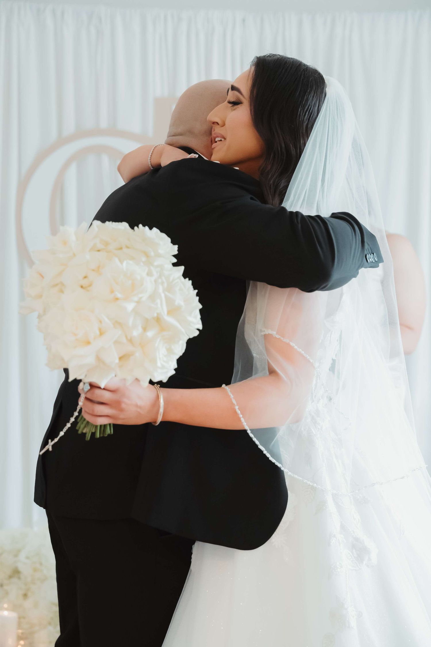 A tender embrace between a bride in white veil and their partner in black suit, with a white bouquet of flowers.