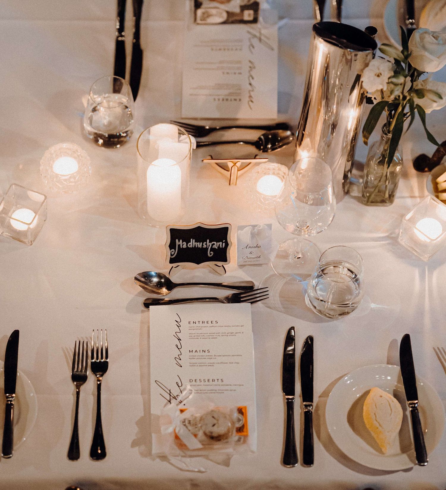 Elegant wedding reception place setting with glowing candles, menu cards, and silver utensils on a white tablecloth.