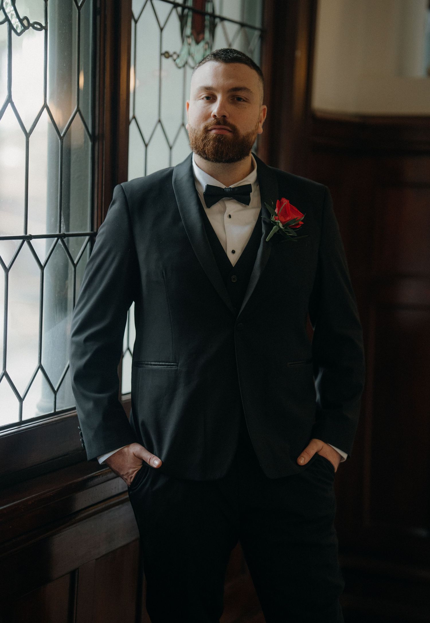 Groom in black tuxedo with bow tie and red rose boutonniere standing by stained glass window.