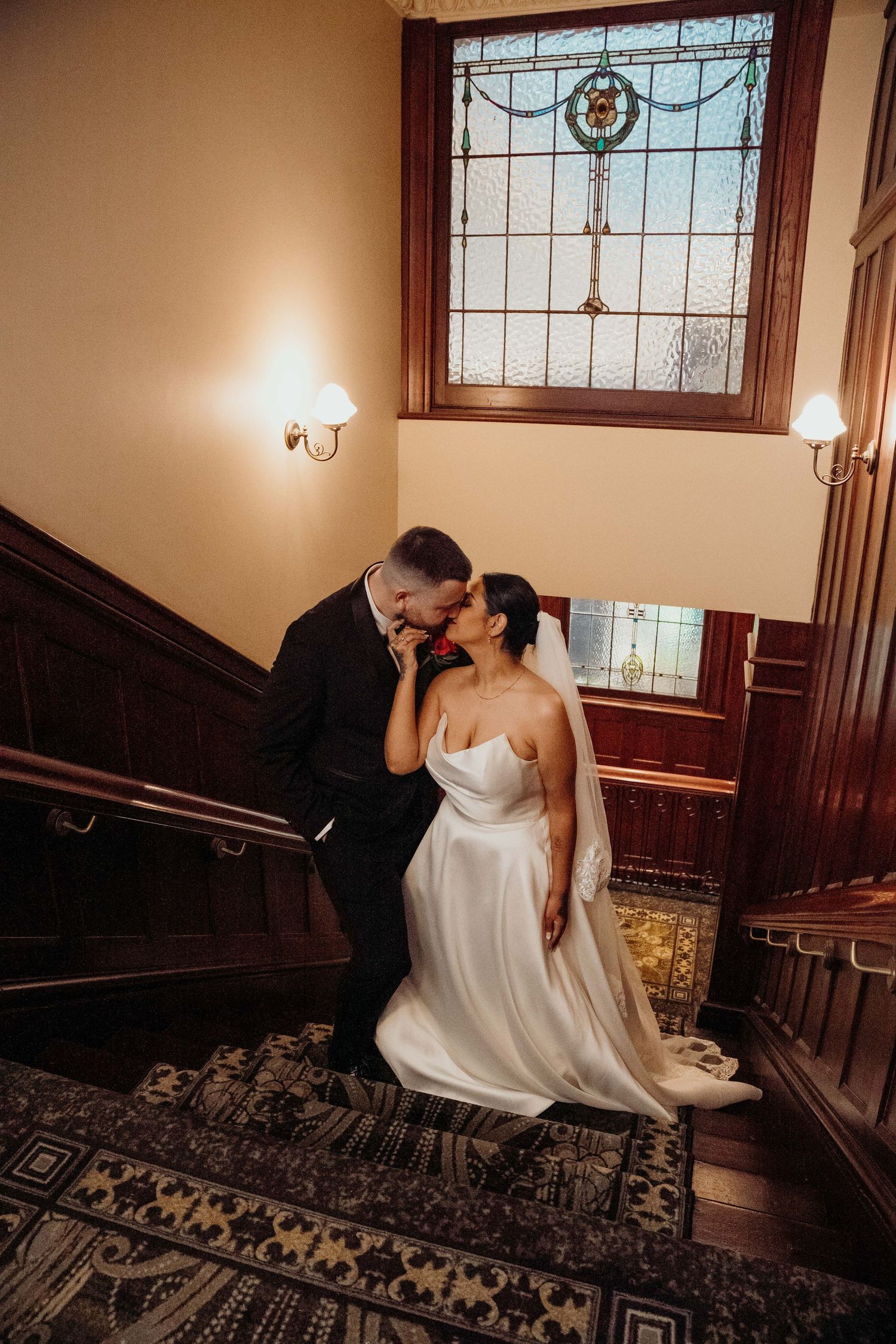 Bride and groom share a romantic kiss on an elegant staircase with stained glass window backdrop.
