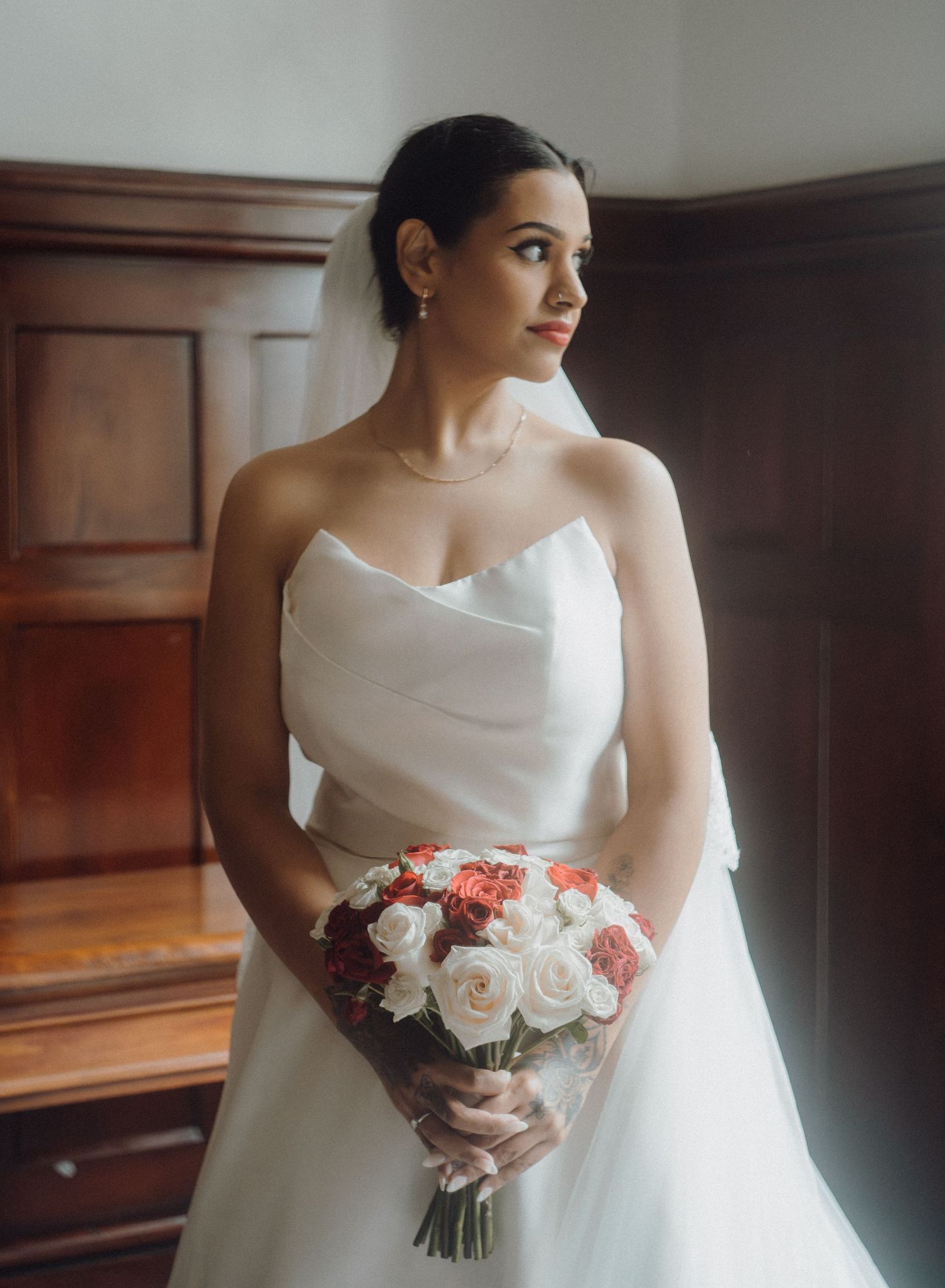 Elegant bride in strapless white gown holding red and white rose bouquet, standing by wooden door with veil.