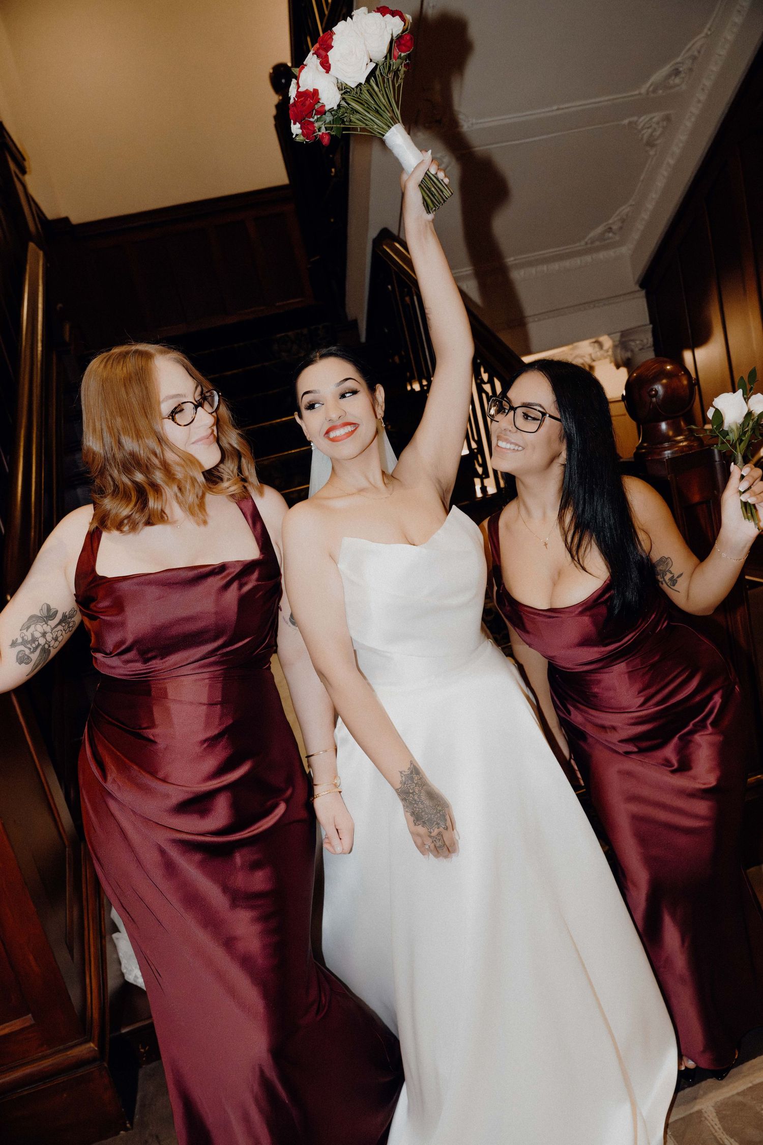 Bride in white gown raises bouquet with two bridesmaids in burgundy satin dresses on staircase.