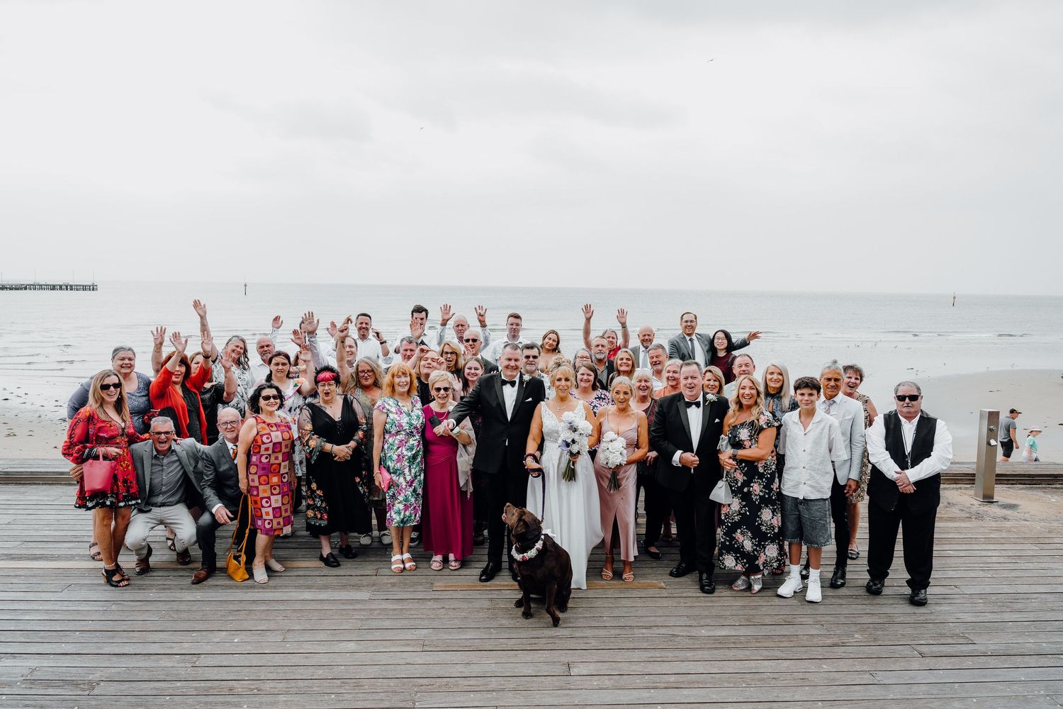 A large wedding party poses together on a wooden boardwalk by the beach.