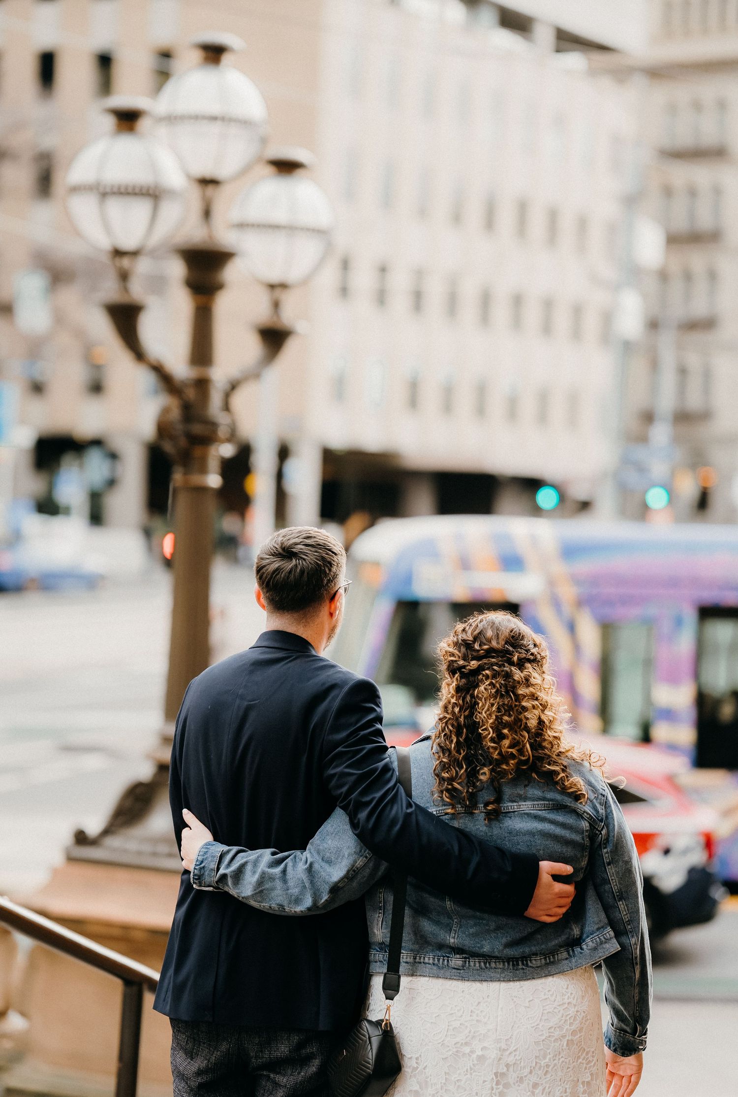 A couple sits together on a stone wall overlooking a European city street with vintage lampposts and buildings.