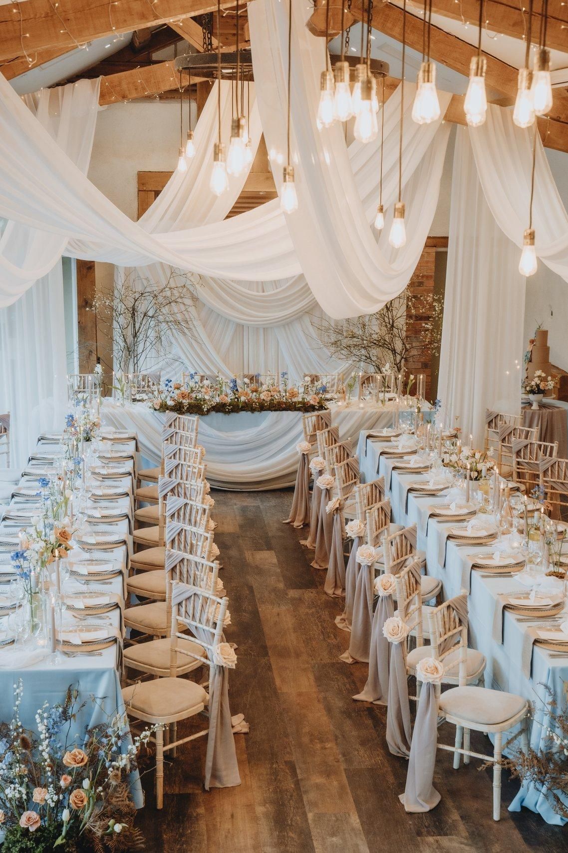 Elegant wedding reception venue with white draped fabric, hanging lights, and long banquet tables set with blue accents.