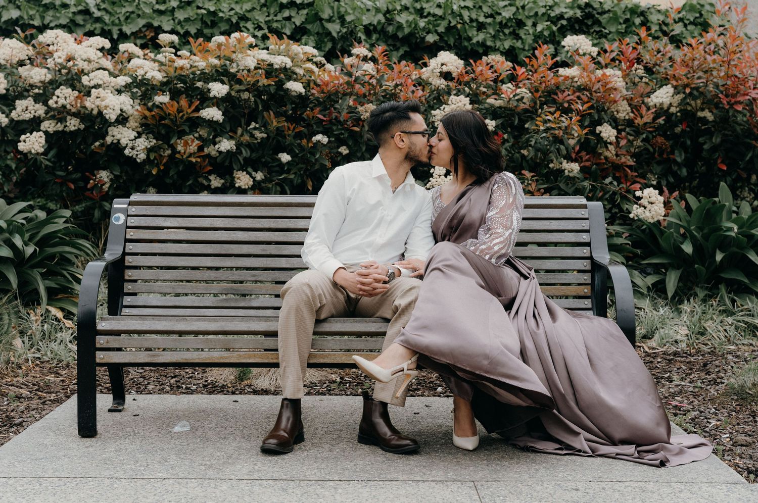 A romantic couple sits together on a wooden bench in a garden with blooming orange flowers in the background.