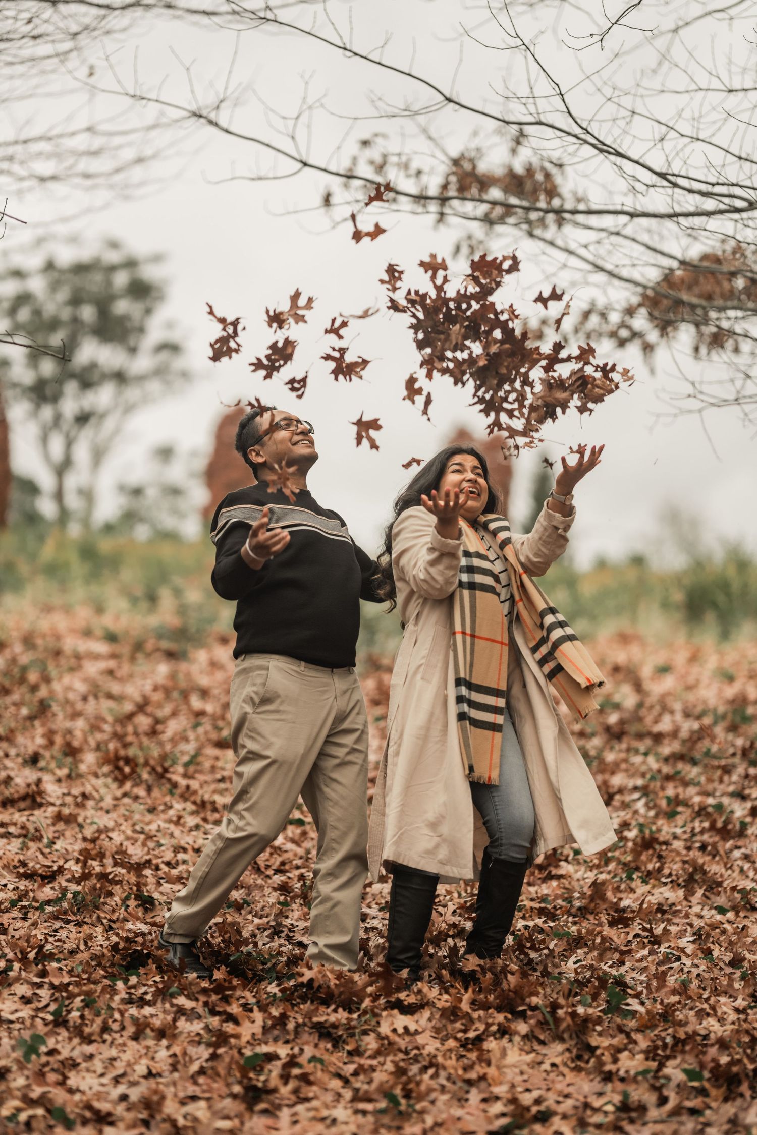 A couple enjoys a playful autumn moment tossing fallen leaves in the air at a park on an overcast day.