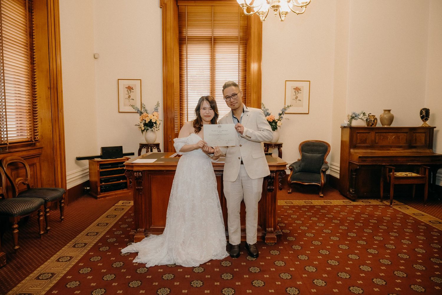 A couple stands together in wedding attire inside an elegant room with vintage furnishings and decor.