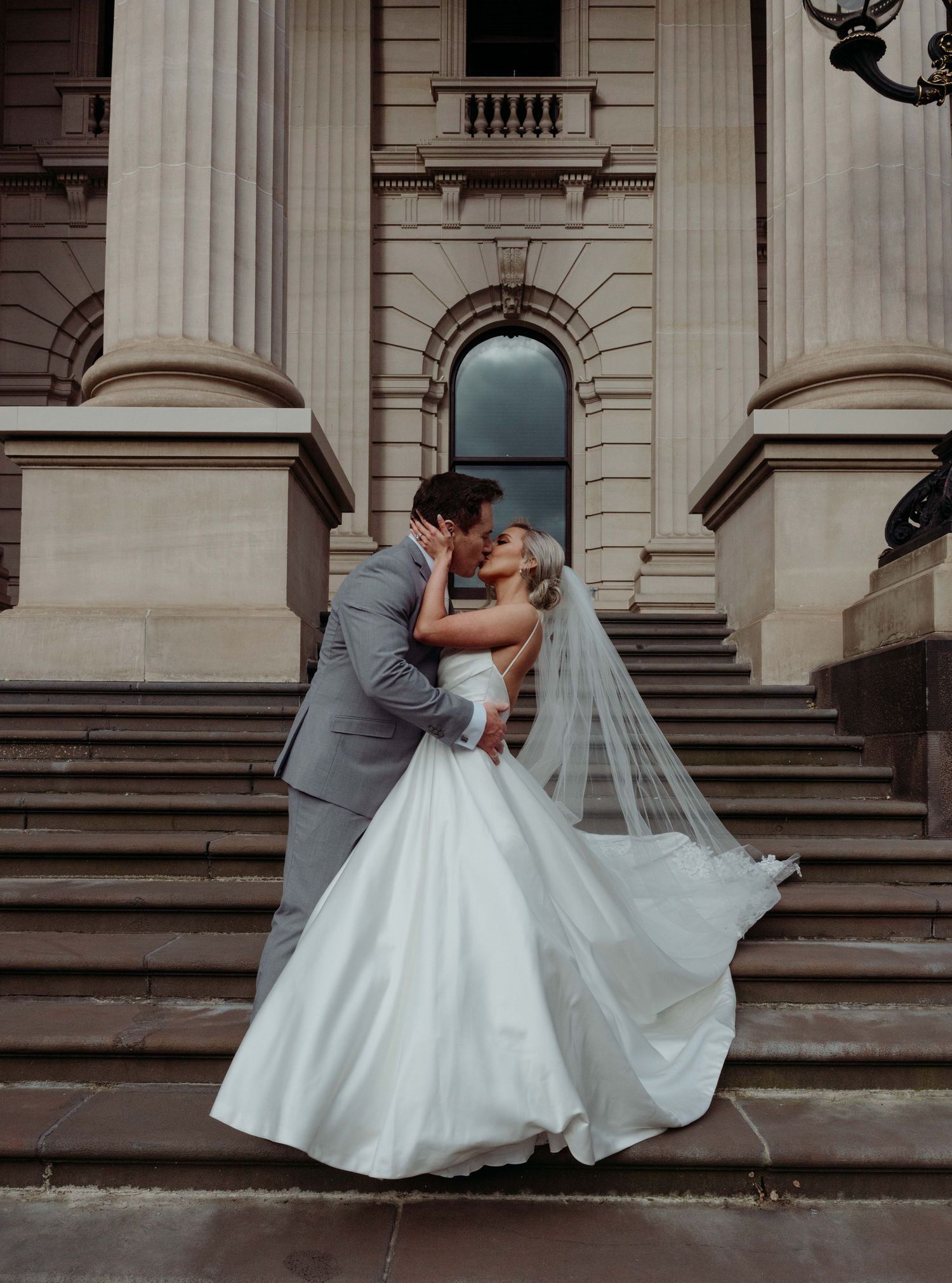 A romantic wedding couple in gray and white attire share a tender embrace on the steps of a grand stone building.